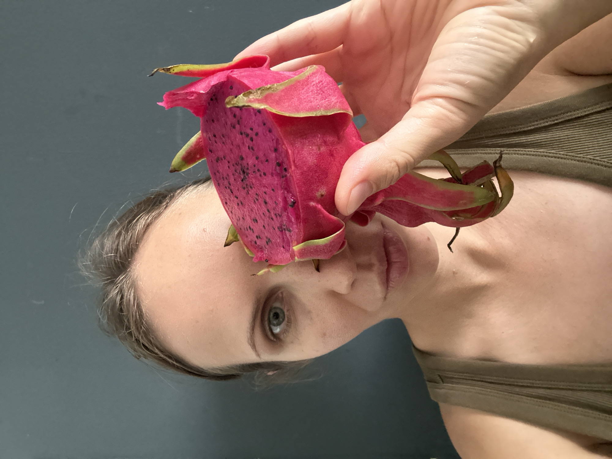 A close-up, medium shot shows a woman in Fethiye, Turkey, holding a slice of pink dragon fruit to her face. The woman's left eye is visible, and her skin is fair. She is wearing a dark green tank top. The background is a plain dark gray wall. The dragon fruit is vibrant pink with small black seeds and green and pink bracts. The woman's hand is gently holding the fruit.