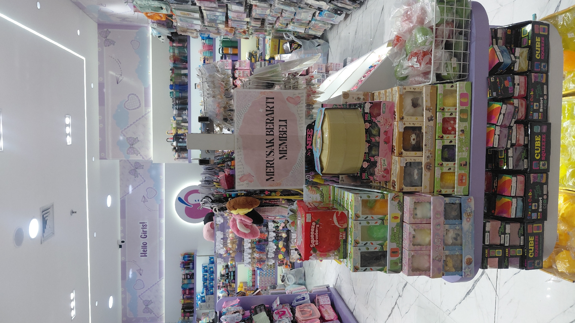 The image shows a retail store interior, likely in Purwokerto, Indonesia, with a focus on toys and novelty items. The store is brightly lit with fluorescent lighting and decorated with pastel colors and cloud motifs. In the foreground, shelves are stacked with various packaged toys, including squishy toys, stress balls, and blind boxes. A prominent display in the center features a large yellow round container and boxes of "Squeeze Strawberry" toys. A sign with text in a foreign language, likely Indonesian, is visible on a stand in front of the display. In the background, shelves are stocked with more merchandise, including water bottles and various accessories. The store appears to be a "Hello Girls!" themed shop, targeting a young female demographic. There are no people clearly visible in the frame.