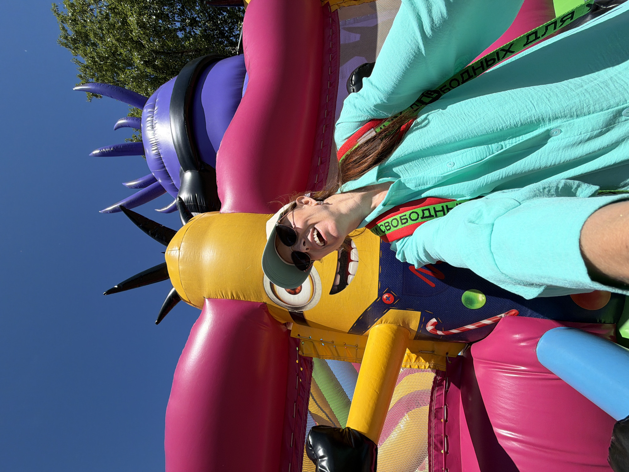 In this selfie, a woman with long brown hair, wearing a light blue long-sleeved shirt, a white baseball cap, and sunglasses, smiles broadly. She is positioned in front of a large, inflatable structure that appears to be a bounce house. The inflatable features a yellow Minion character from the Despicable Me franchise, complete with a glove extended outwards. Behind the Minion, there's a large pink and purple inflatable element with spikes, against a clear blue sky. The woman has a red and green strap across her chest. The scene suggests an outdoor setting, likely a park or event space, with trees visible in the background. The inscription on the strap is "СВОБОДНЫХ ДЛЯ".