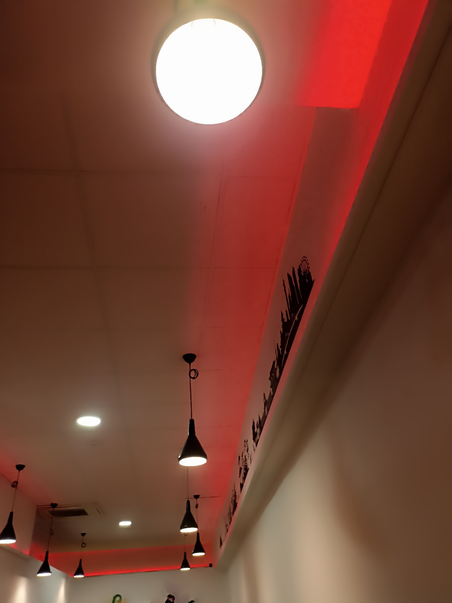 The image shows a ceiling with multiple pendant lights and recessed lighting. A red LED strip light runs along the edge of the ceiling, illuminating a black silhouette of a city skyline with a Ferris wheel. The room appears to be indoors and dimly lit, with the red light casting a warm glow. There are no people or clear interactions visible. The setting is a commercial space, possibly a restaurant or bar, given the decorative lighting and skyline motif. The location in Galway, Ireland, is not discernible from the visual information provided in the image.