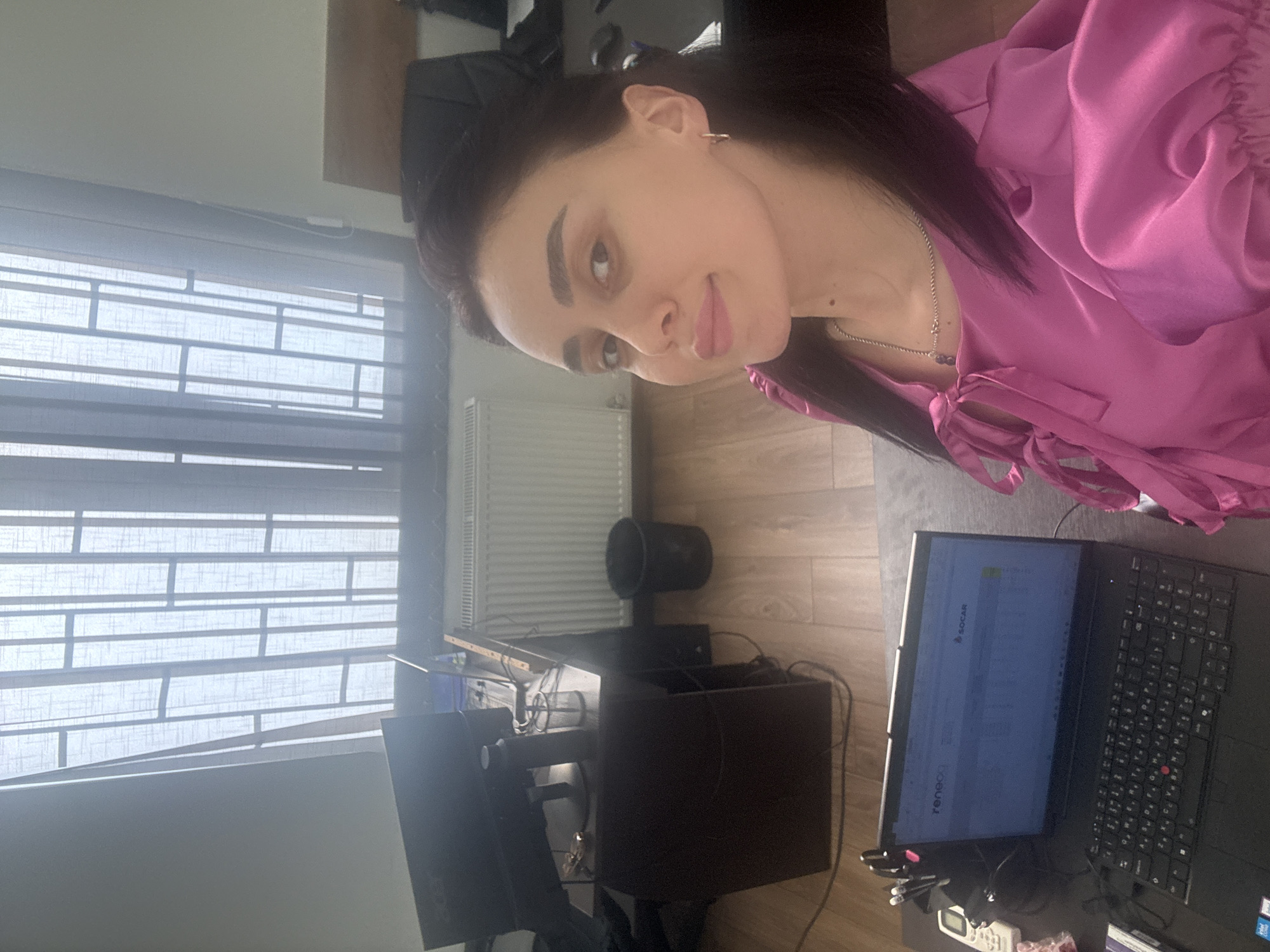 A woman with dark hair and light skin is sitting at a desk, wearing a magenta blouse and a necklace. She is looking at the camera. On the desk is a laptop with the words "SOCAR" and "renecO" visible on the screen, along with office supplies and other computer equipment. A window with blinds is visible behind the desk, and a radiator is visible to the side. The room appears to be an office space. The image provides no verifiable information to confirm it was taken in Tbilisi, Georgia.