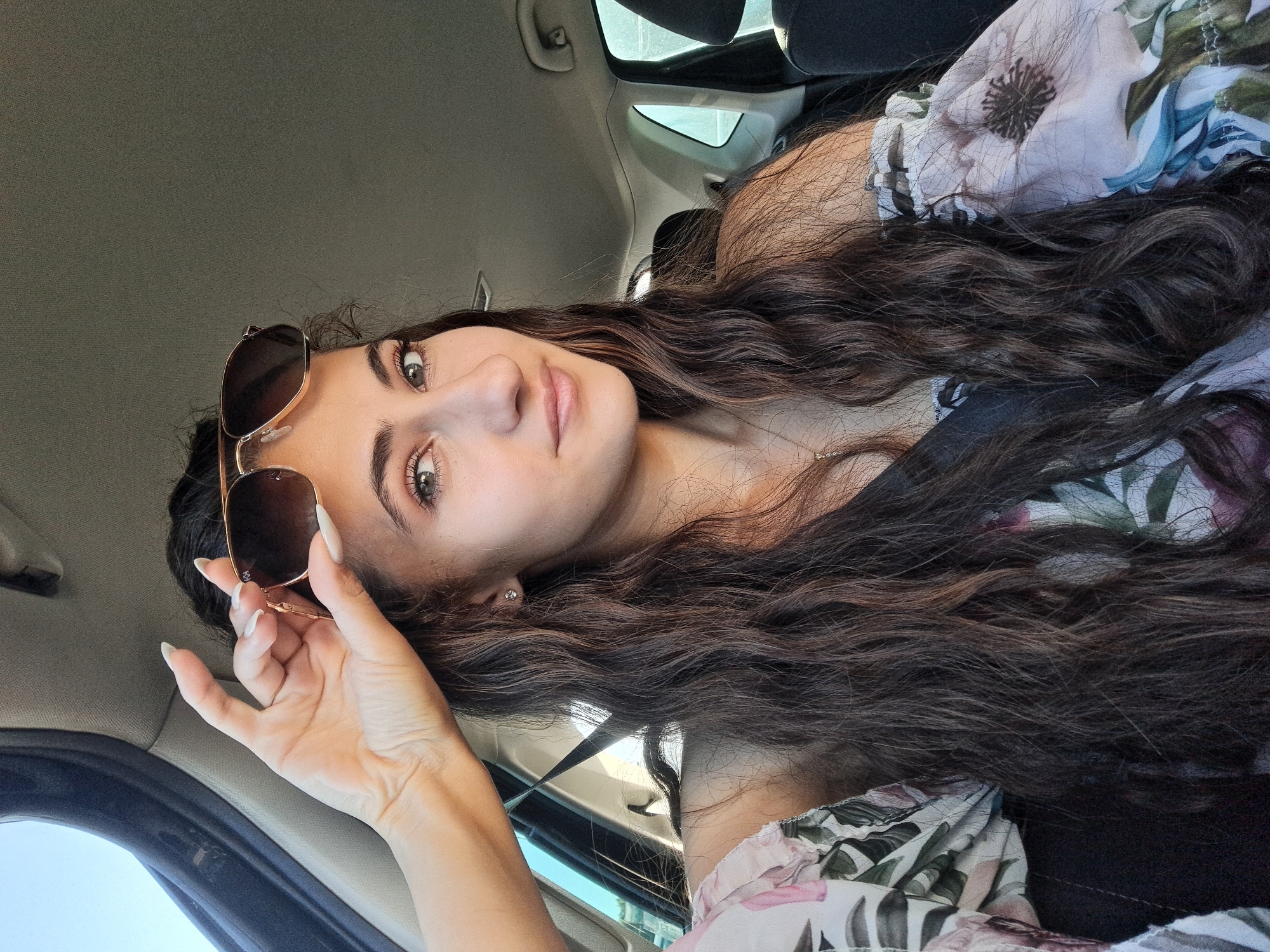 A young woman with long, wavy brown hair and green eyes is taking a selfie in the passenger seat of a car. She is wearing sunglasses on top of her head and a floral-print off-the-shoulder top. Her left hand is raised, holding the sunglasses in place. The interior of the car is visible, including the headrest of the driver's seat and the car's ceiling. The lighting suggests it is daytime, with sunlight filtering into the car. The background is out of focus, showing the exterior of the car and some foliage.