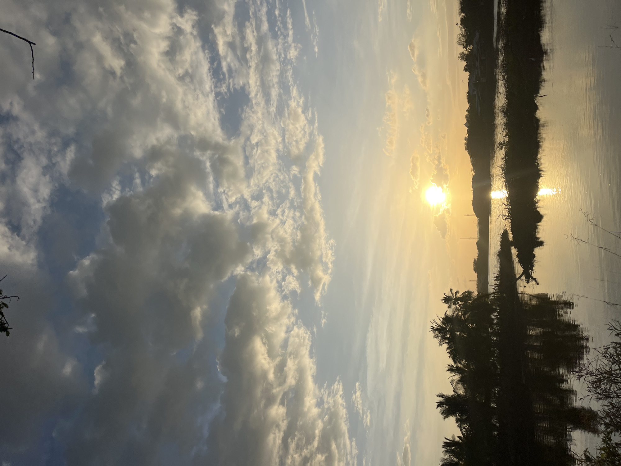 The image shows a sunset over a body of water, possibly a lake or river. The sun is bright, positioned near the horizon, casting a golden glow across the sky and reflecting on the water. The sky is filled with clouds of varying sizes and densities. The right side of the image features silhouetted trees and vegetation along the water's edge, their reflections visible in the water. The location is possibly in Lhokseumawe, Indonesia, as requested. There are no people visible in the frame.