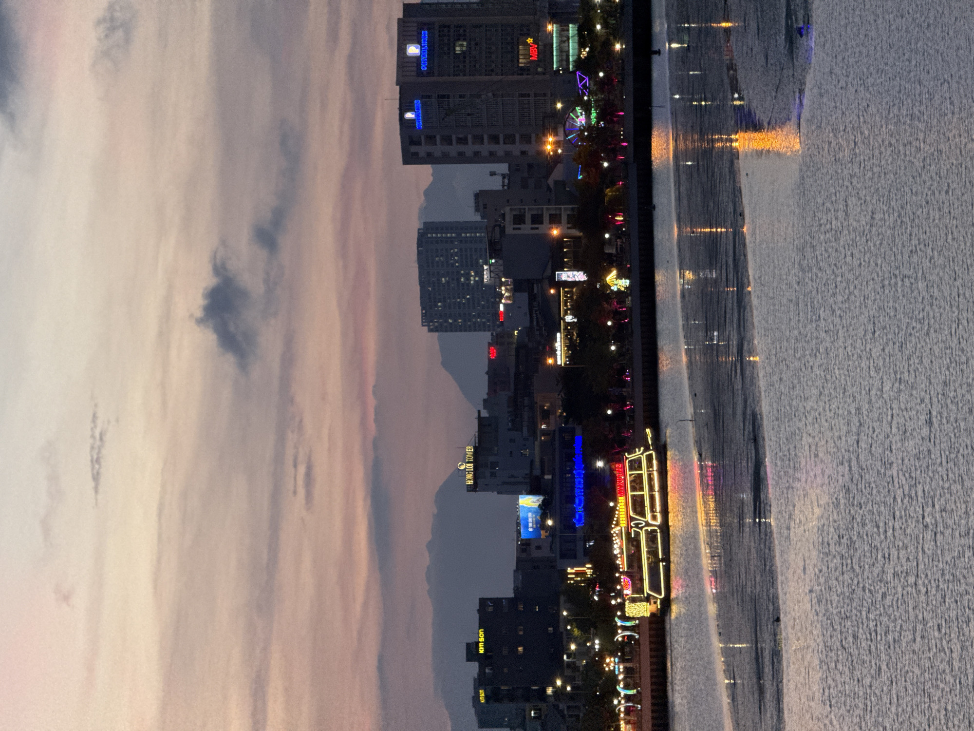 The image depicts a twilight scene of a city skyline along a body of water, likely a river or bay, in Da Nang, Vietnam. The sky displays a gradient of soft pinks and grays, indicating the transition from day to night. Wispy clouds are visible, adding texture to the sky.
The urban landscape features several multi-story buildings, some of which are illuminated with neon signs and lights. One building prominently displays the text "HỒNG LĨNH TOWER." Another building has text that appears to be "Kim Son." Lights from the buildings reflect on the rippling surface of the water, creating a shimmering effect.
Along the waterfront, there are decorative lights and what appears to be a boat or a structure resembling a boat, also adorned with colorful lights, suggesting a festive or entertainment area. The overall atmosphere is calm and serene, with the city lights creating a vibrant contrast against the darkening sky. The time of day is clearly evening or dusk.