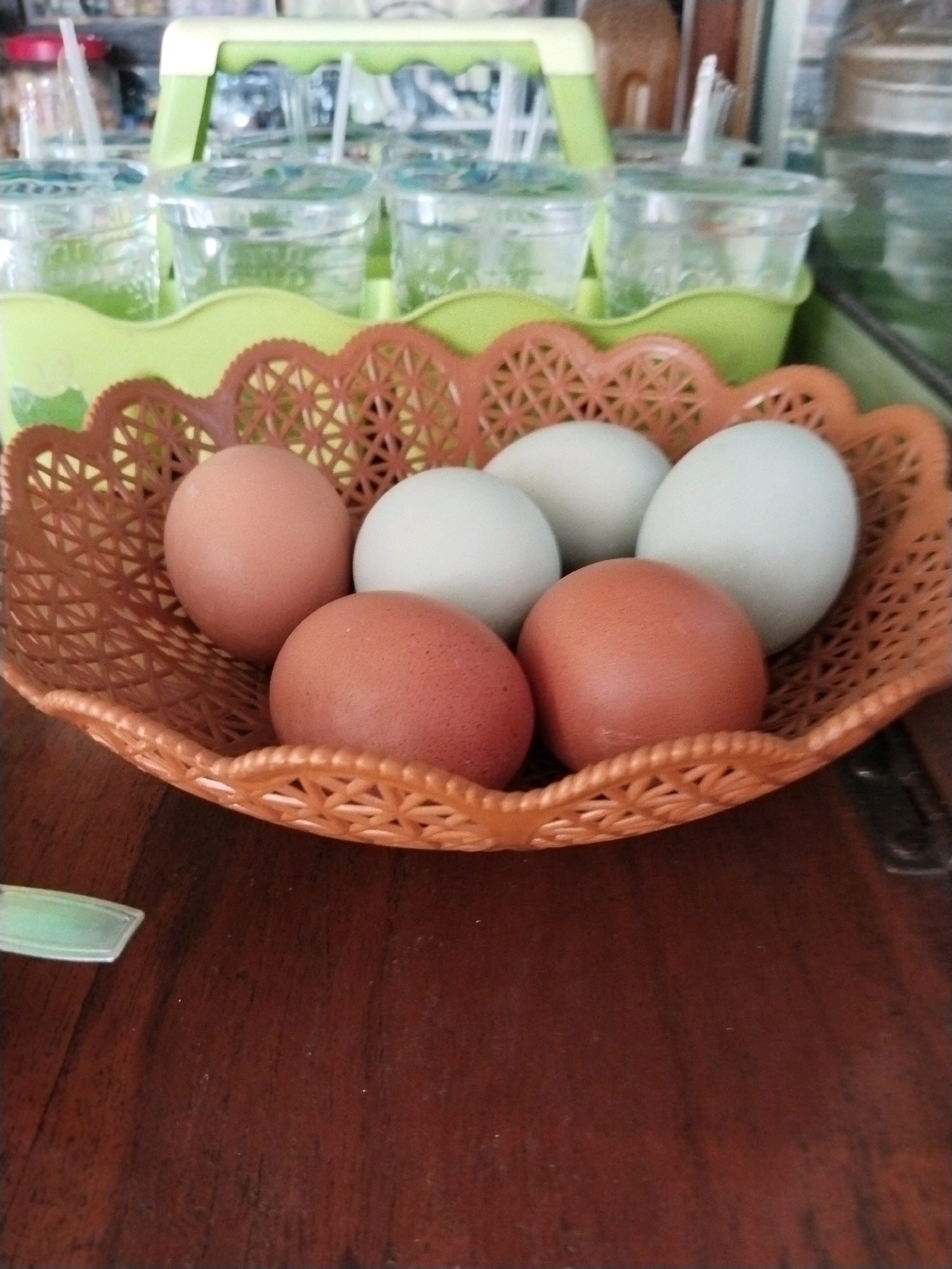 The image shows a close-up view of a woven orange basket filled with six eggs. Three of the eggs are a reddish-brown color, and the other three are a pale blue-green. The basket is sitting on a dark brown wooden surface, possibly a table. In the background, out of focus, there is a green plastic tray holding several clear plastic cups with straws, suggesting a beverage setup. The lighting is moderately bright, casting soft shadows. The location in the city of Trenggalek, Indonesia cannot be determined from the image.