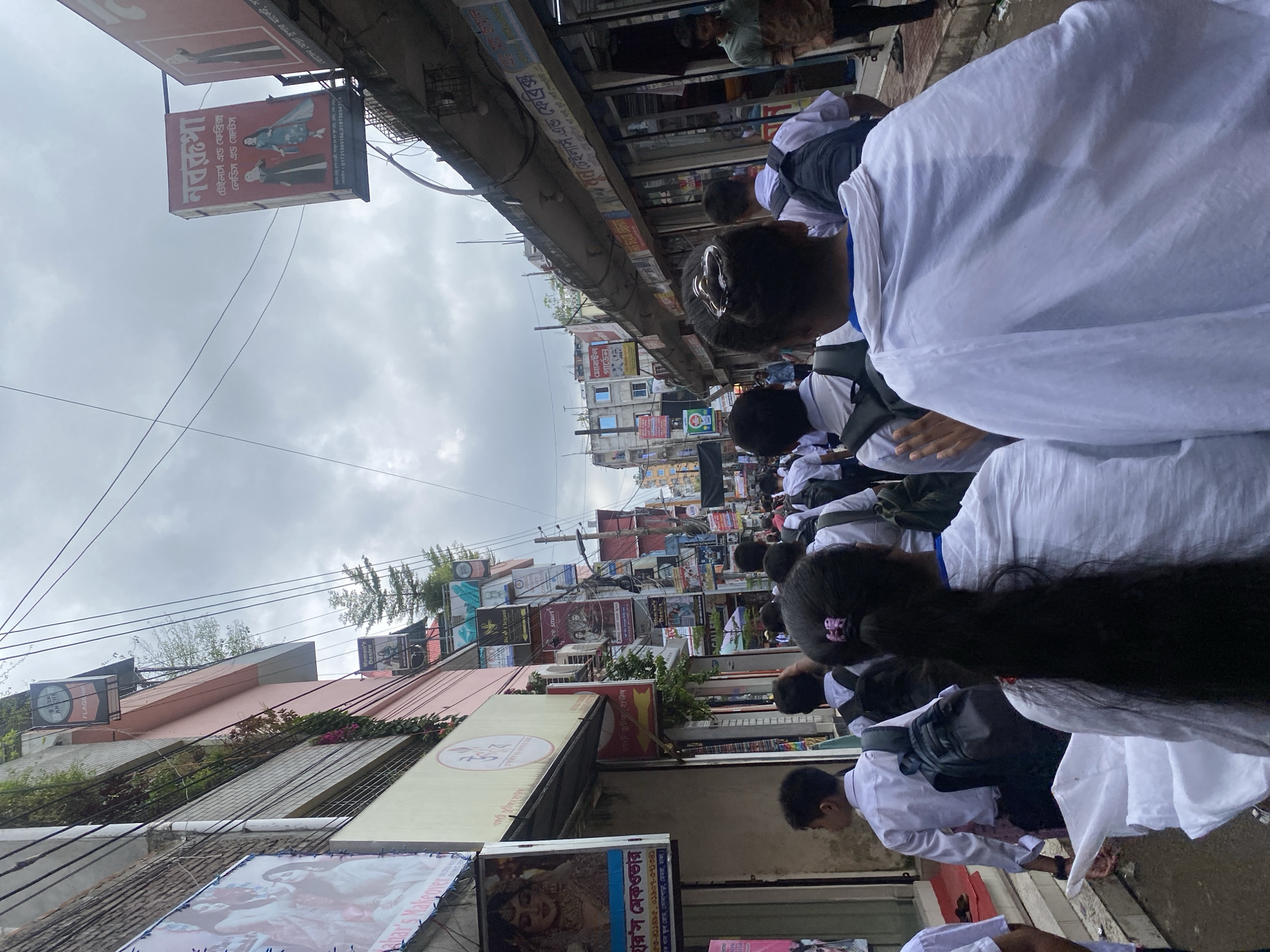 This is a street-level, medium-shot, eye-level, outdoor photograph taken in a bustling market area in Dhaka, Bangladesh. The scene depicts a narrow street lined with multi-story buildings housing various shops and businesses. Numerous signs, predominantly in Bengali, are displayed on the building facades, indicating the nature of the establishments. Several people are walking along the street, with a group of young individuals, presumably students wearing white shirts and some carrying backpacks, walking away from the camera. The sky is overcast and cloudy, suggesting a diffused light condition. The overall impression is one of a busy commercial district with pedestrian traffic.
