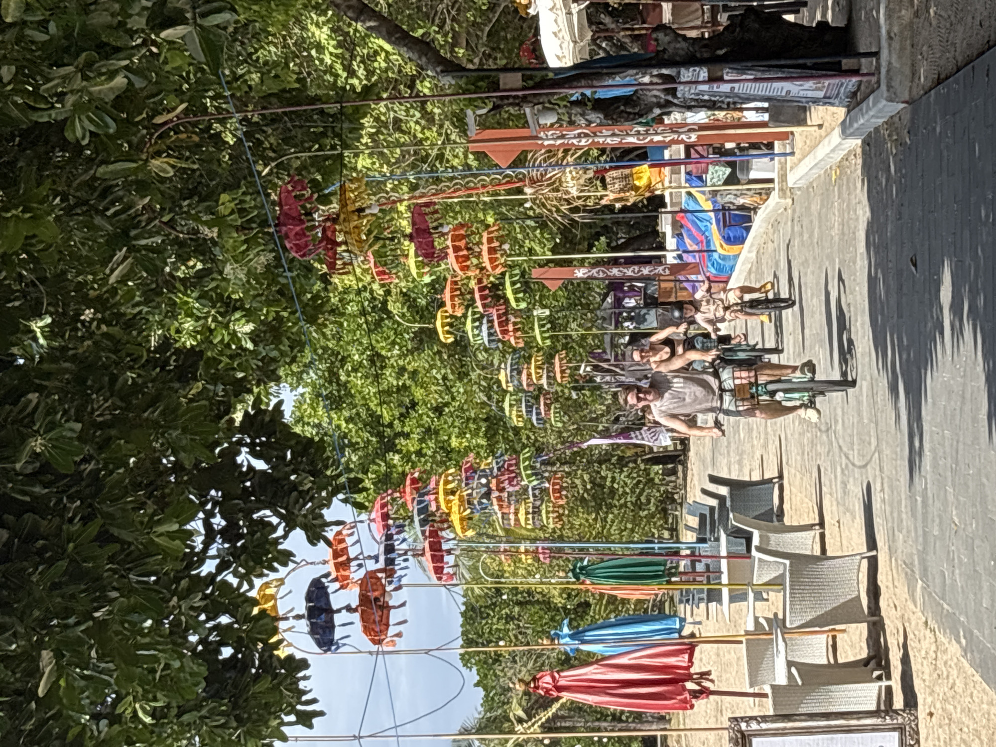 In Kuta Selatan, Indonesia, a family is seen cycling on a paved walkway alongside a sandy beach. Overhead, a multitude of colorful, ornate umbrellas are suspended from wires, creating a festive canopy. In the background, the lush green foliage of trees lines the area, with further decorative elements and beachside establishments visible. The foreground shows several white wicker tables and chairs set up on the sand, suggesting a dining or relaxation area. The family, consisting of two adults and a child, are all on bicycles, with the adults wearing casual clothing and the child wearing a helmet. The scene captures a lively, tropical atmosphere.