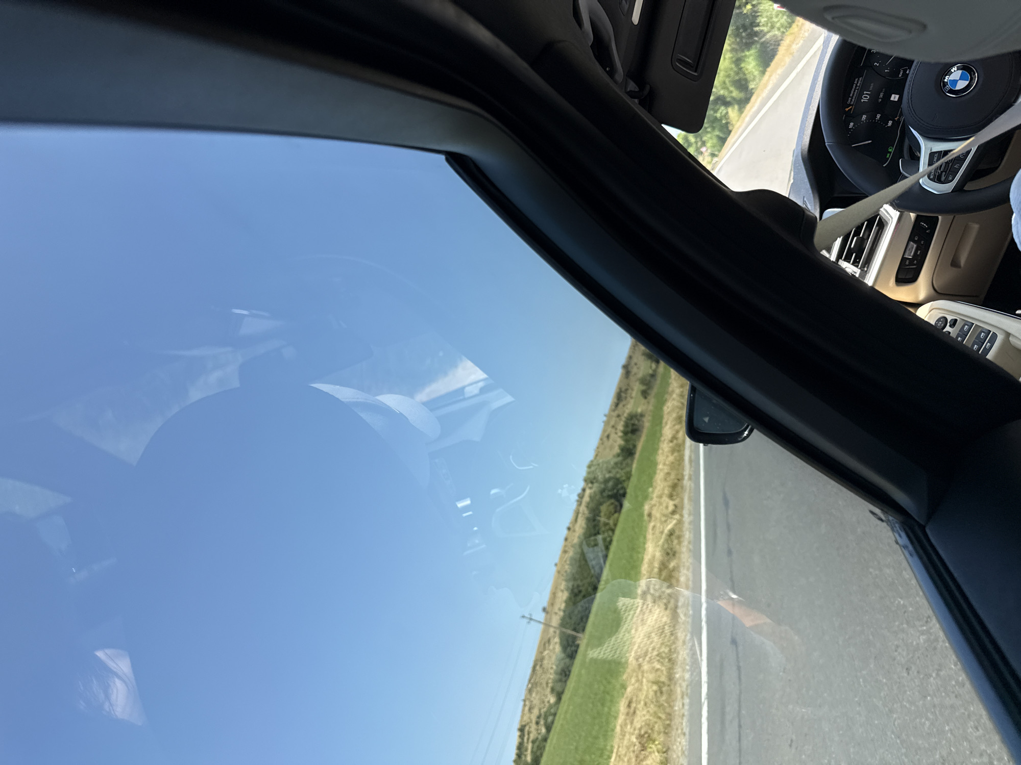 The image shows the interior of a car with a clear sky visible through the sunroof. A person's head and shoulder are reflected in the sunroof, obscuring some of the sky. The car appears to be in motion, as indicated by the blurred road and passing landscape. The landscape consists of green fields and rolling hills, with some trees and utility poles visible. The interior of the car includes a dashboard with a digital display, a steering wheel, and a seatbelt. The overall impression is one of a scenic drive on a sunny day. The specific location in the city of Dumbrăvița, Romania, cannot be determined from the image alone, as there are no identifying landmarks or signs.