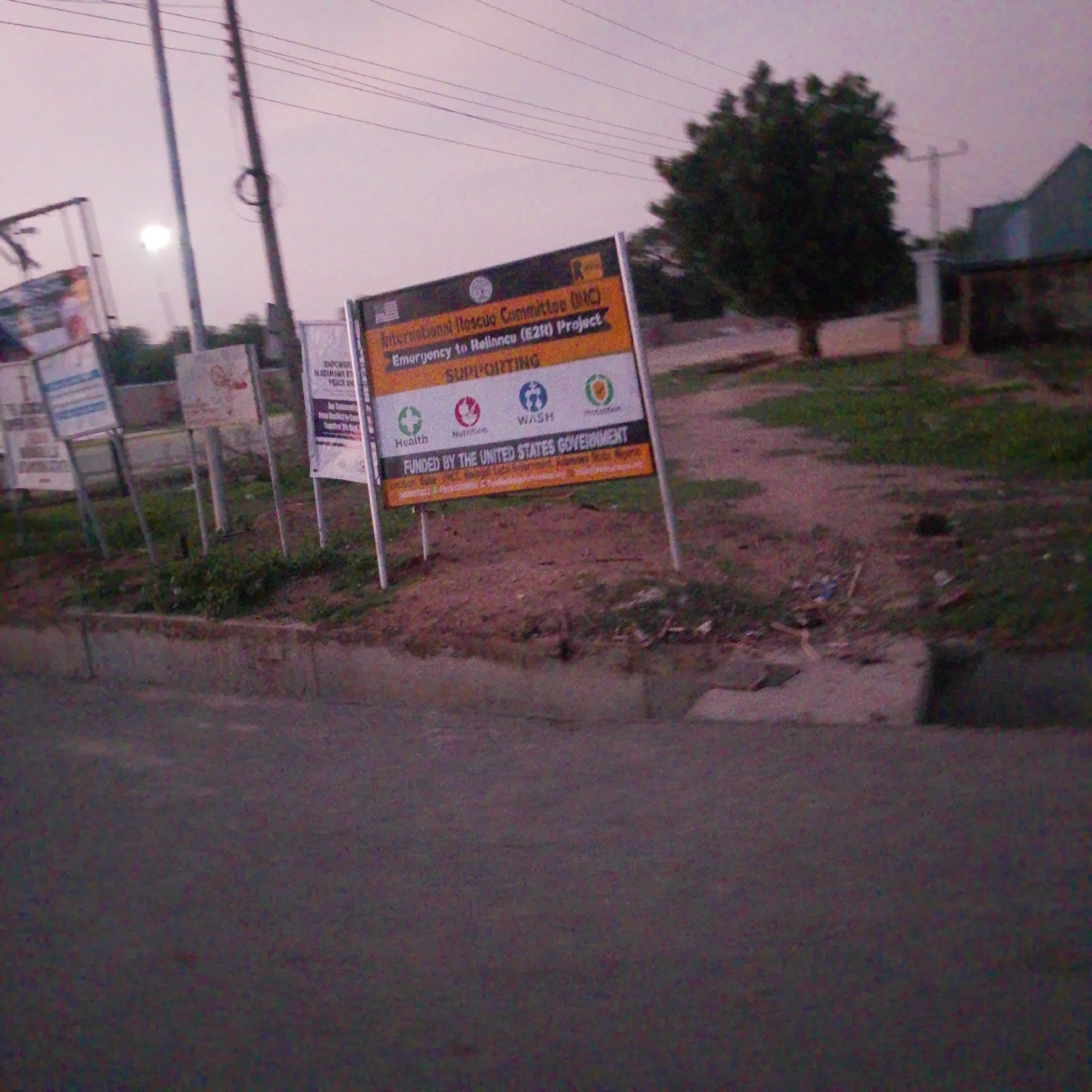 In Wuro Allahamdu, Nigeria, this image captures a street scene featuring a prominent sign for the International Rescue Committee (IRC) Emergency to Resilience (E2R) Project. The sign indicates support for Health, Nutrition, WASH (Water, Sanitation, and Hygiene), and Protection. The project is funded by the United States Government. In the background, a large tree and a building are visible, along with utility poles and overhead wires. The ground is covered with dirt and some scattered debris. The sky is overcast, suggesting twilight or a cloudy day. Other signs are also present in the background, though less legible.