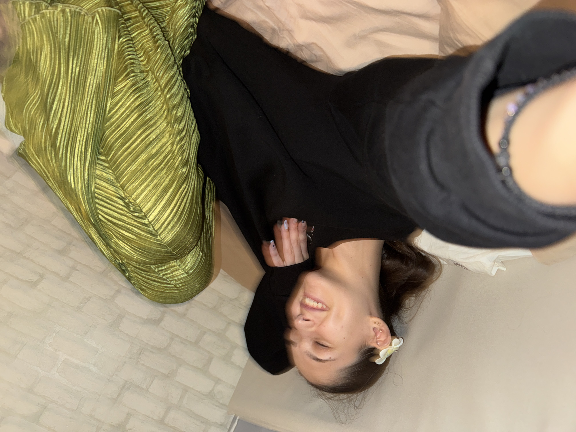 A young woman with brown hair is lying on her back on a surface that appears to be a bed or couch. She is wearing a black long-sleeved top and has a white flower tucked behind her left ear. Her eyes are closed, and she is smiling. Her right hand is resting on her head, and her left hand is extended. She is wearing a beaded bracelet on her left wrist. To her upper right, a piece of draped, shiny green fabric is visible, which appears to be clothing or upholstery. The background wall has a textured, brick-like pattern. The overall scene suggests a relaxed and casual setting, possibly in a bedroom or living room. There is no indication of the city of Sofia, Bulgaria, in the image.