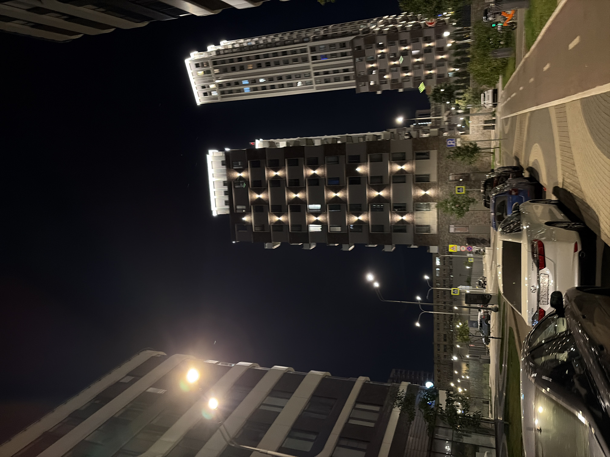 The image depicts a nighttime urban scene in Moscow, Russia, specifically within a modern residential complex. Several multi-story buildings, some with illuminated windows and architectural lighting, dominate the skyline. In the foreground, various cars are parked along a street or in a parking area. Streetlights illuminate the paved surfaces, including what appears to be a pedestrian path or bicycle lane. The environment is characterized by clean lines, contemporary architecture, and a well-maintained urban landscape. No people are visible in the image.