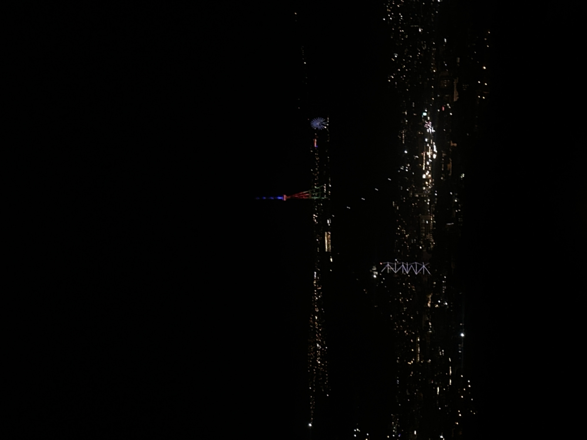 The image depicts a nighttime scene, with the right side showing an illuminated cityscape and the left side being predominantly dark. In the city, there's a Ferris wheel visible, and what appears to be a lit-up tower structure. The lights from the city are reflecting off of surfaces, creating bright, specular highlights. A white sign of the word "NMNMK" is seen.