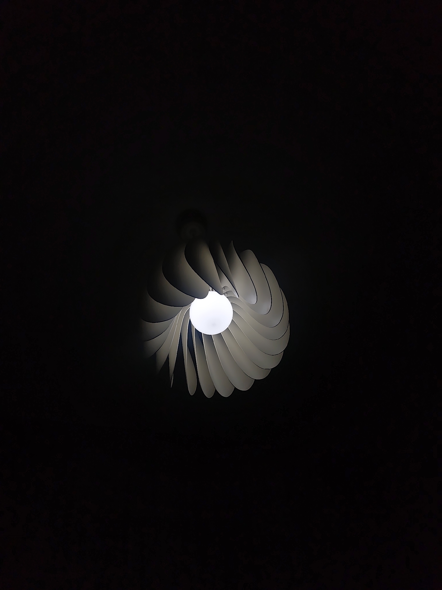 The image features a modern light fixture hanging from a dark ceiling. The fixture has a round lightbulb surrounded by white, petal-like structures arranged in a spiral. The lighting is dim, with most of the scene obscured by darkness. The exact location in Mosomishte, Bulgaria cannot be determined from the visual information provided.