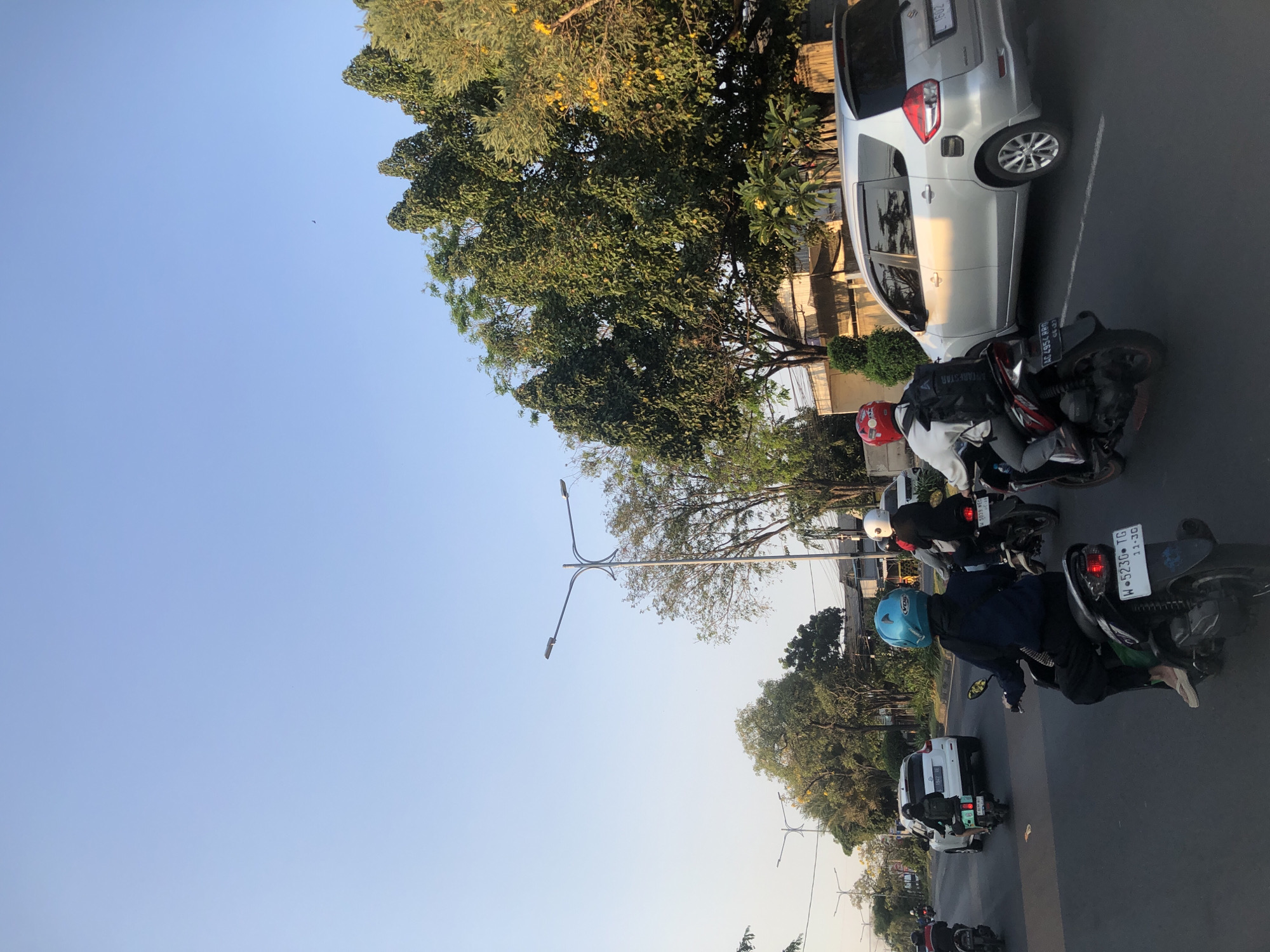 The image depicts a street scene in Gelam, Indonesia, during daylight. The sky is clear and a pale blue. Several motorcycles are on the road, with riders wearing helmets. A silver minivan is also visible, driving on the road. Trees line the street, with some bearing yellow flowers. Streetlights are present along the road. The overall impression is a typical urban environment with moderate traffic.