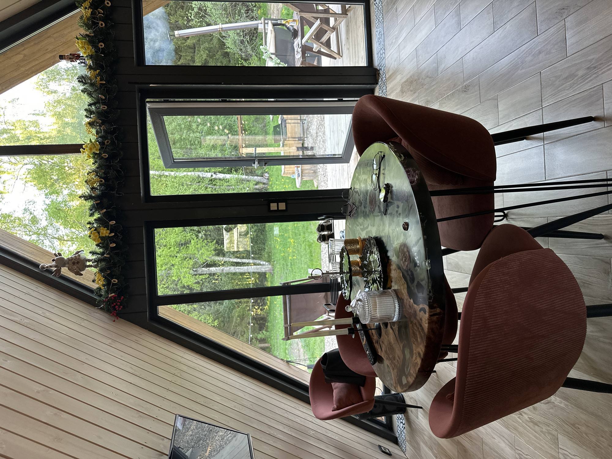 A residential interior in Малеевка, Russia, showcases a dining space. A round wooden table, adorned with glassware and serving pieces, is surrounded by four maroon-colored upholstered chairs with black metal legs. A wide window behind the table offers an outdoor view of trees and grass. The window frame is decorated with greenery and ornaments. Interior walls are lined with light-colored wooden paneling. Floor tiles are light grey. Through the open door a wood burning hot tub can be seen outside.