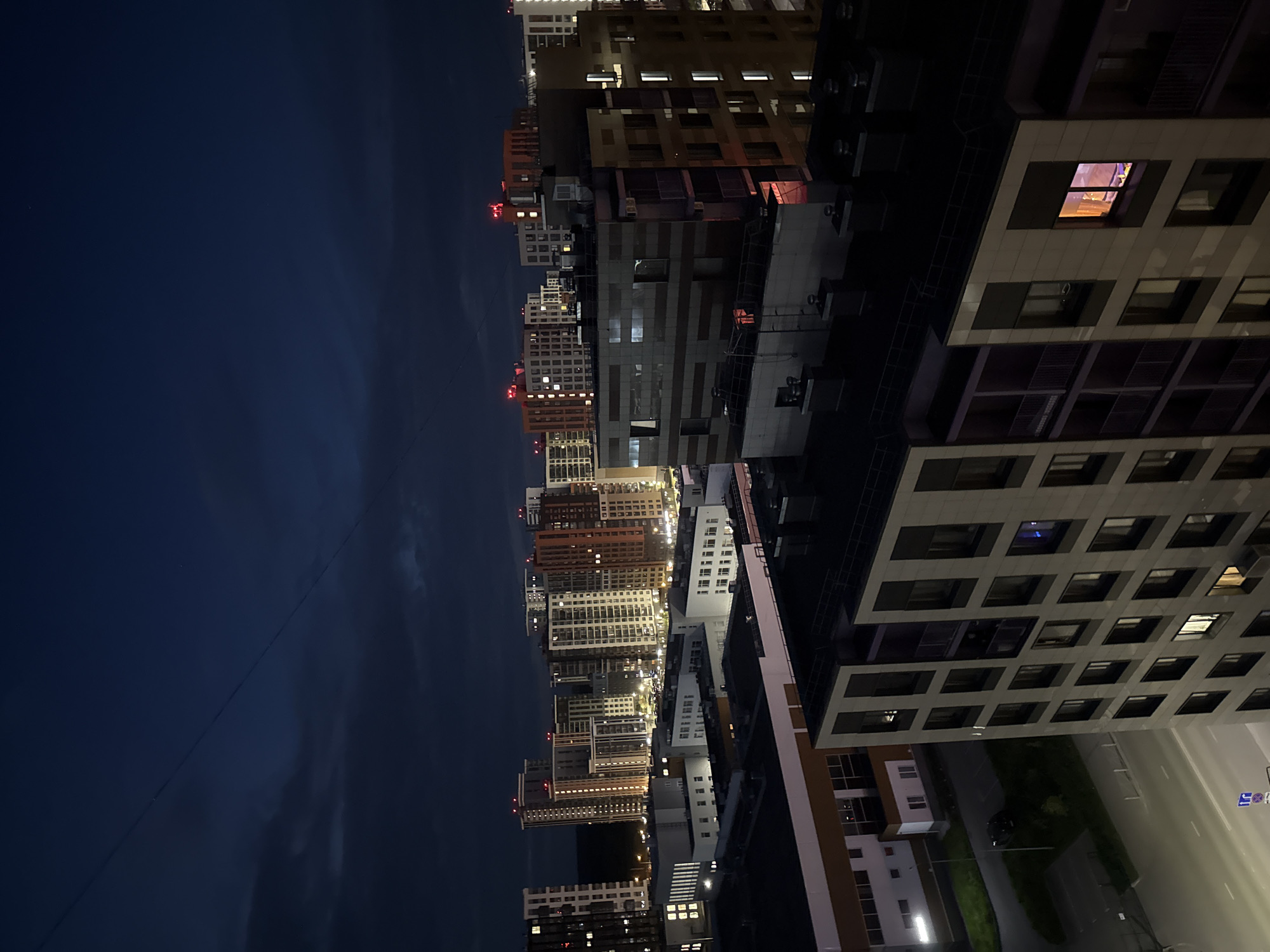 The image shows a nighttime aerial view of a modern residential complex in the city of Perm, Russia. The sky is dark blue with some clouds, and a power line is visible stretching diagonally across the upper portion of the frame. In the foreground, there are several multi-story apartment buildings with illuminated windows. Some buildings have balconies with railings, and the facades are constructed from various materials, including concrete and what appears to be decorative paneling. The street level is visible in the lower left, with a car parked on the side and some lampposts providing light. The overall scene depicts a typical urban residential area at night, with the warm glow of interior lights contrasting with the dark exterior.