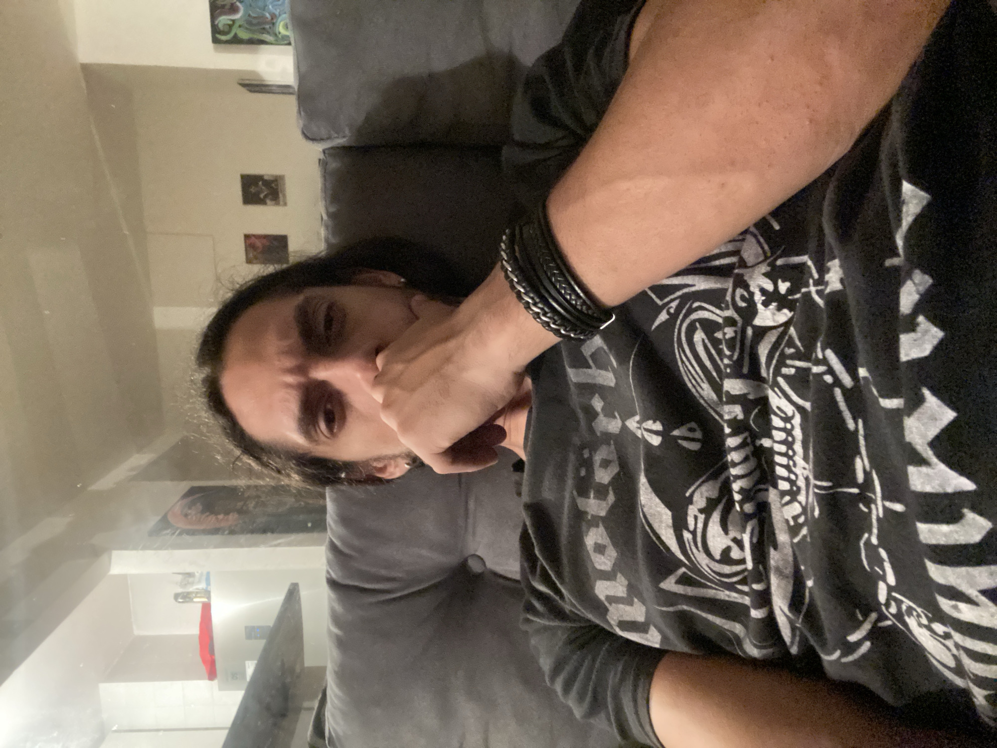 An indoor portrait-style photograph shows a person, likely male, with long dark hair, dark eyes, and a pensive expression. The subject is captured in a somewhat horizontal orientation, appearing to be lying down, with their gaze directed towards the camera. One hand is partially covering their mouth and chin area, contributing to the thoughtful look.
The person is wearing a dark, possibly black, t-shirt adorned with intricate white graphic designs, which appear to include stylized text or symbols that are not clearly legible. A stack of multiple bracelets, made from both leather and metallic chain, is visible on their right wrist. They are resting on what appears to be a gray sofa or a large cushion.
In the background, on a light-colored wall, several small framed pictures or artworks can be seen. A prominent feature of the scene is a large, reflective surface on the left side, possibly glass or a mirror. This reflection reveals parts of another adjacent room, showing white walls, what might be a doorway, a red object, and some metallic fixtures. The lighting suggests an interior setting, possibly during the evening. The scene is located in Buenos Aires, Argentina.