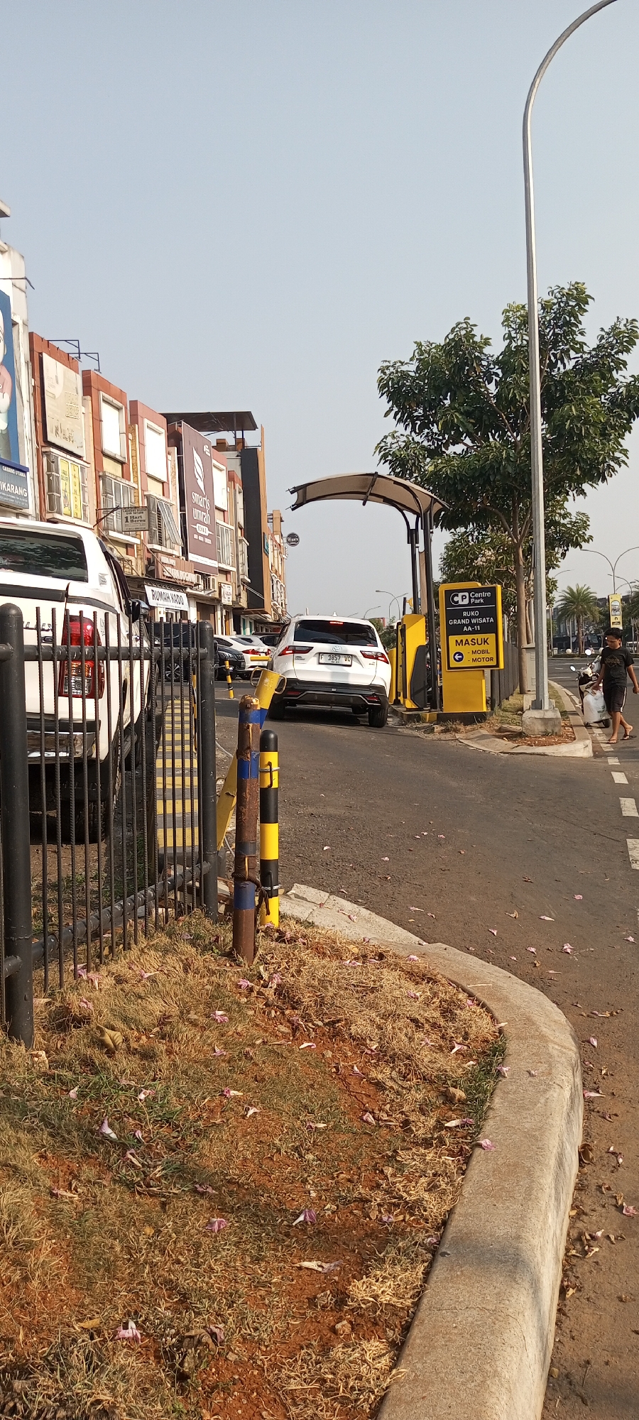 In Lambangjaya, Indonesia, a white SUV is exiting a gated area. The vehicle, a Toyota RAV4 with license plate F 3857 JC, is positioned on a paved road. To the right of the SUV, a yellow ticket booth with signage indicating "RUKO GRAND WISATA AA-11 MASUK - MOBIL - MOTOR" is visible. Behind the booth, a person in a black t-shirt and shorts walks away from a parked motorcycle. A tall street lamp with a curved arm is on the right side of the image, and a tree with green leaves is situated near it. In the background, a row of buildings with multiple stories and various advertisements is seen. A black metal fence with vertical bars and yellow diagonal stripes runs along the left side of the road, separating it from a patch of dry grass with scattered pink flowers.