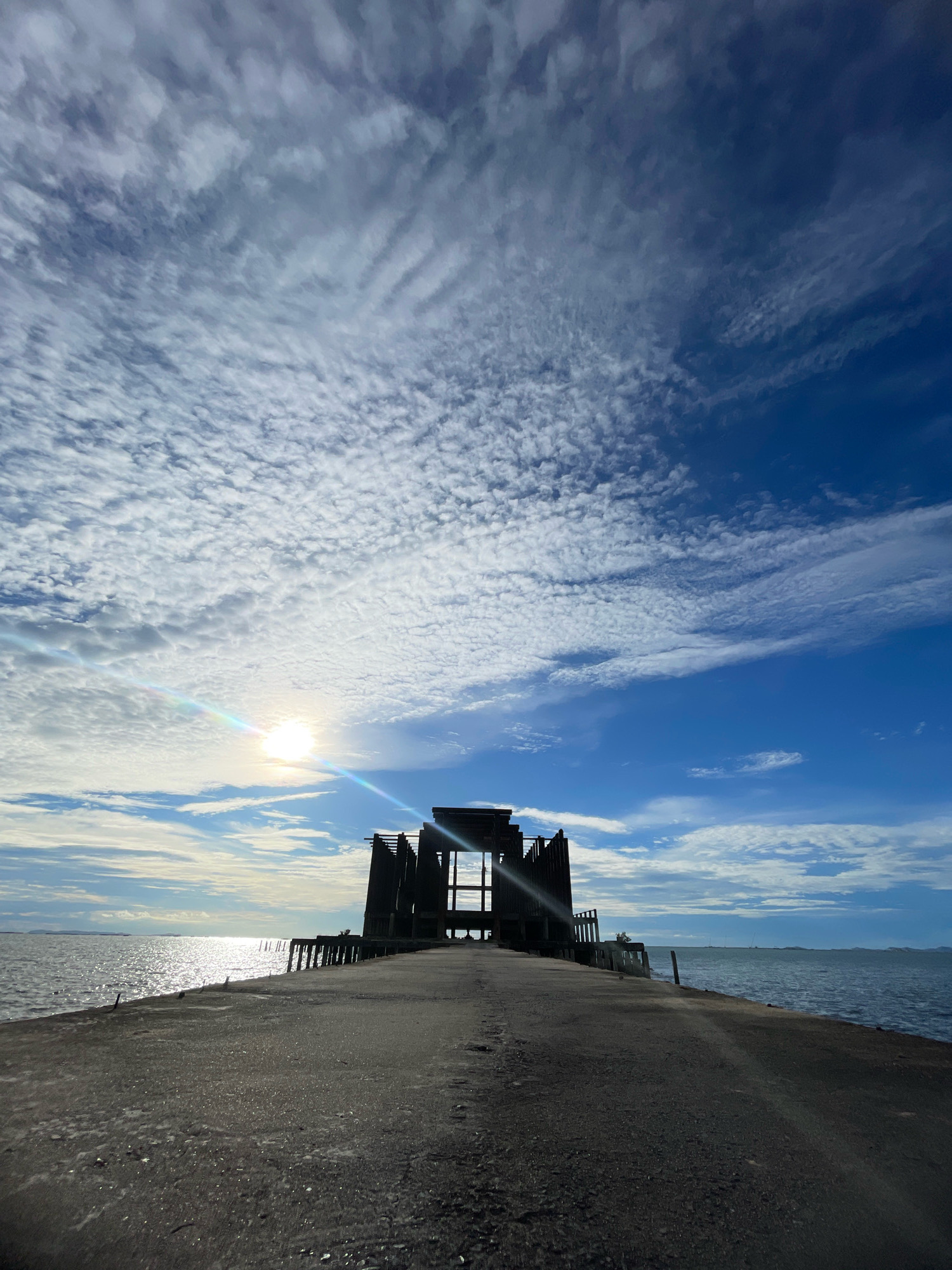 The image shows a concrete pier extending into the sea. The pier leads towards a silhouetted, unfinished structure made of metal and wood, which appears to be an abandoned building or a construction site. The sky is a vibrant blue with scattered white clouds, and the sun is visible, casting a bright glare on the water. The sea is calm, with gentle waves lapping at the pier. In the distance, a hazy coastline is visible. The overall scene suggests a sense of desolation and decay, juxtaposed with the beauty of the natural environment. This location is likely part of the coastline of Pattaya City, Thailand, known for its beaches and urban development.