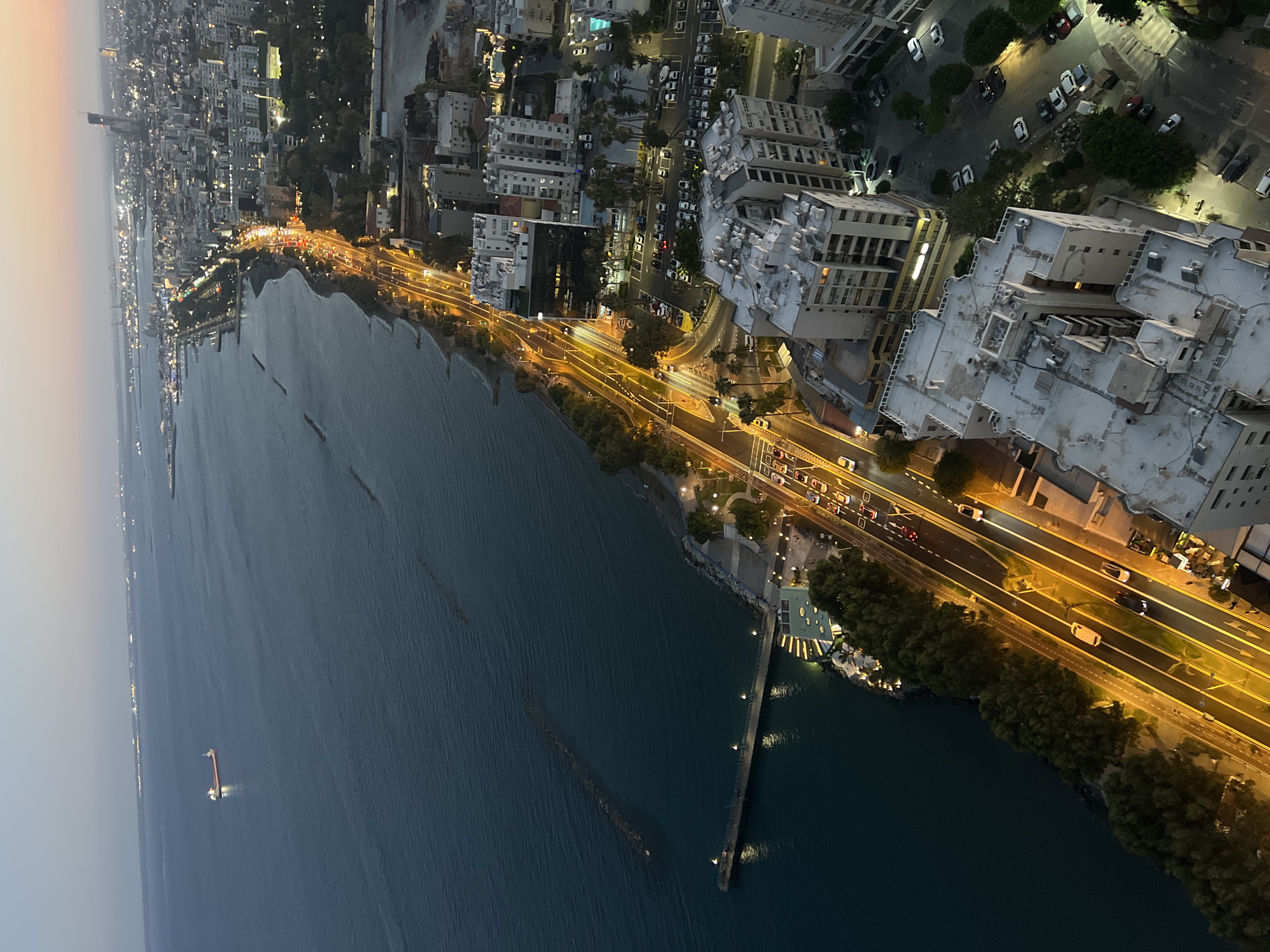 This is an aerial, high-angle, wide shot of a coastal city at dusk. The scene captures a bustling cityscape adjacent to a calm, dark blue sea. A prominent multi-lane road runs along the coastline, illuminated by bright streetlights, with numerous cars creating streaks of light due to the long exposure, indicating traffic and movement. Buildings of varying heights and architectural styles, many with lit windows, line the road and extend inland, creating a dense urban environment. Green trees and foliage are interspersed among the buildings, softening the urban landscape.
The sea is a significant element, with a long pier extending from the shore, also illuminated by lights. In the distance, a small boat or structure with lights is visible on the water. The sky transitions from a pale orange-pink near the horizon, suggesting sunset, to a darker, dusky blue overhead. The overall atmosphere is one of urban vibrancy and coastal beauty during the evening transition. No people are clearly discernible as distinct subjects, and no visible text is present in the image. The location is identified as Limassol, Cyprus, which is consistent with the visual cues of a Mediterranean coastal city.