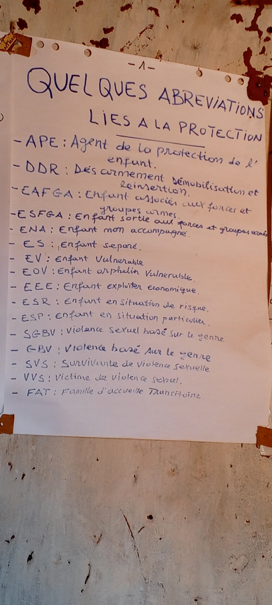 The image shows a sheet of paper pinned to a wall. The paper contains a list of French abbreviations related to the protection of children, likely in an area where French is spoken. The title reads "Quelques abbreviations lies a la protection", which translates to "Some abbreviations related to protection". The abbreviations and their meanings, hand-written in blue ink, are mostly related to children in vulnerable situations (e.g., child soldiers, orphans, exploited children, victims of violence). The wall appears old and possibly damaged. Given the content of the list (child protection, disarmament), the location (Ngalula, DRC) and the language (French), it is possible the paper is related to a humanitarian organization operating in the area.