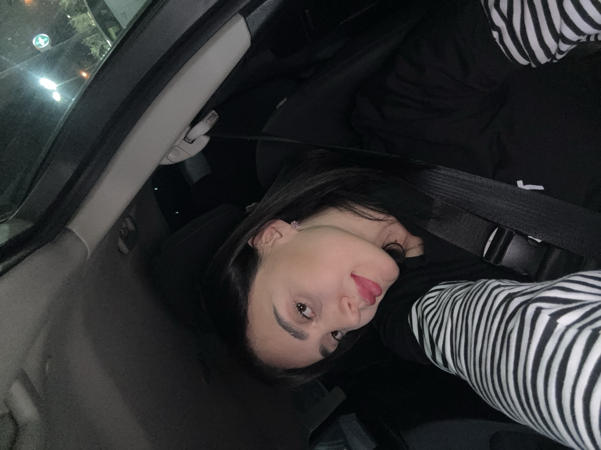 A woman is in the passenger seat of a car. She is wearing a black and white striped long-sleeved shirt. She has dark hair and is wearing a seatbelt. The car's interior is visible, including the A-pillar and the headliner. Outside the window, there are streetlights and a sign with a green cross, which is commonly used to denote a pharmacy. The scene appears to be at night, possibly in Tbilisi, Georgia, based on the potential presence of a pharmacy sign, but without further context, location confirmation is not possible.