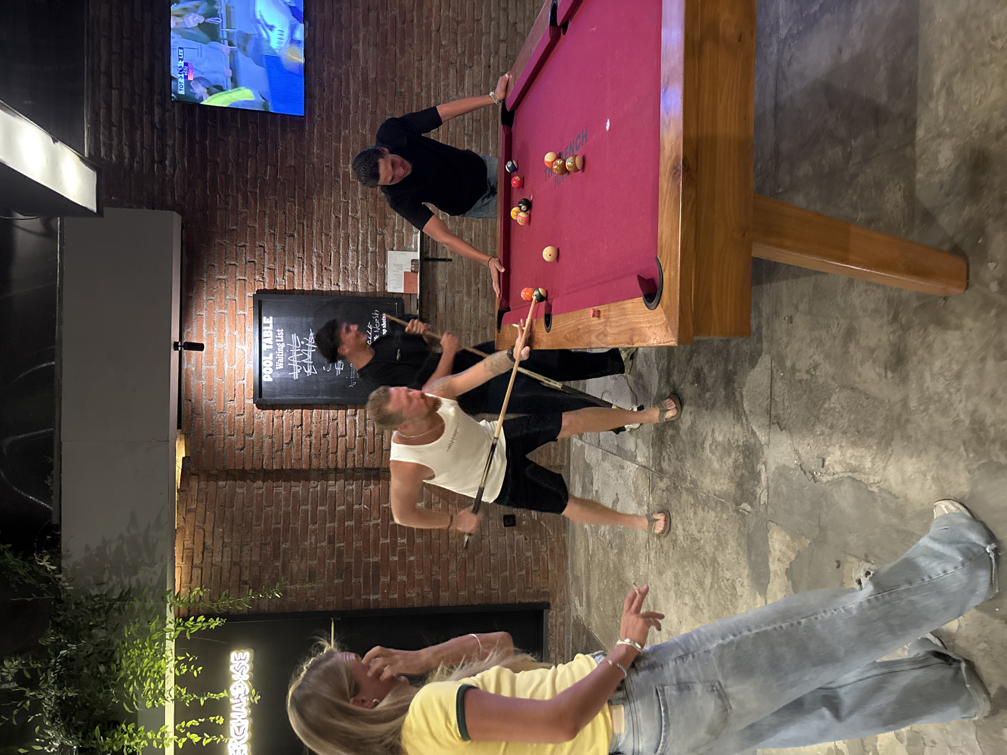 In Kuta Selatan, Indonesia, a group of people is playing pool indoors. A man in a white tank top and black shorts is in the middle of taking a shot, holding a pool cue. Behind him, another man in a black shirt is also holding a pool cue. To the right, a man in a black shirt is leaning over the pool table. The pool table has a red felt surface and several pool balls scattered on it. A woman in a yellow shirt and jeans is standing in the foreground, looking down. A chalkboard with "POOL TABLE Waiting List" written on it is visible on the wall behind the players. A television is mounted on the wall above the chalkboard. The environment suggests a casual setting, possibly a bar or entertainment venue.