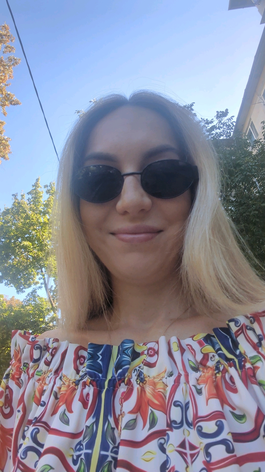 A woman with blonde hair and sunglasses is looking at the camera. She is wearing an off-the-shoulder top with a colorful floral print and a gold necklace with a cross pendant. The background is a clear blue sky with some trees and a building. The image appears to be taken outdoors on a sunny day. The specific location within Chișinău, Moldova cannot be determined from the provided image.