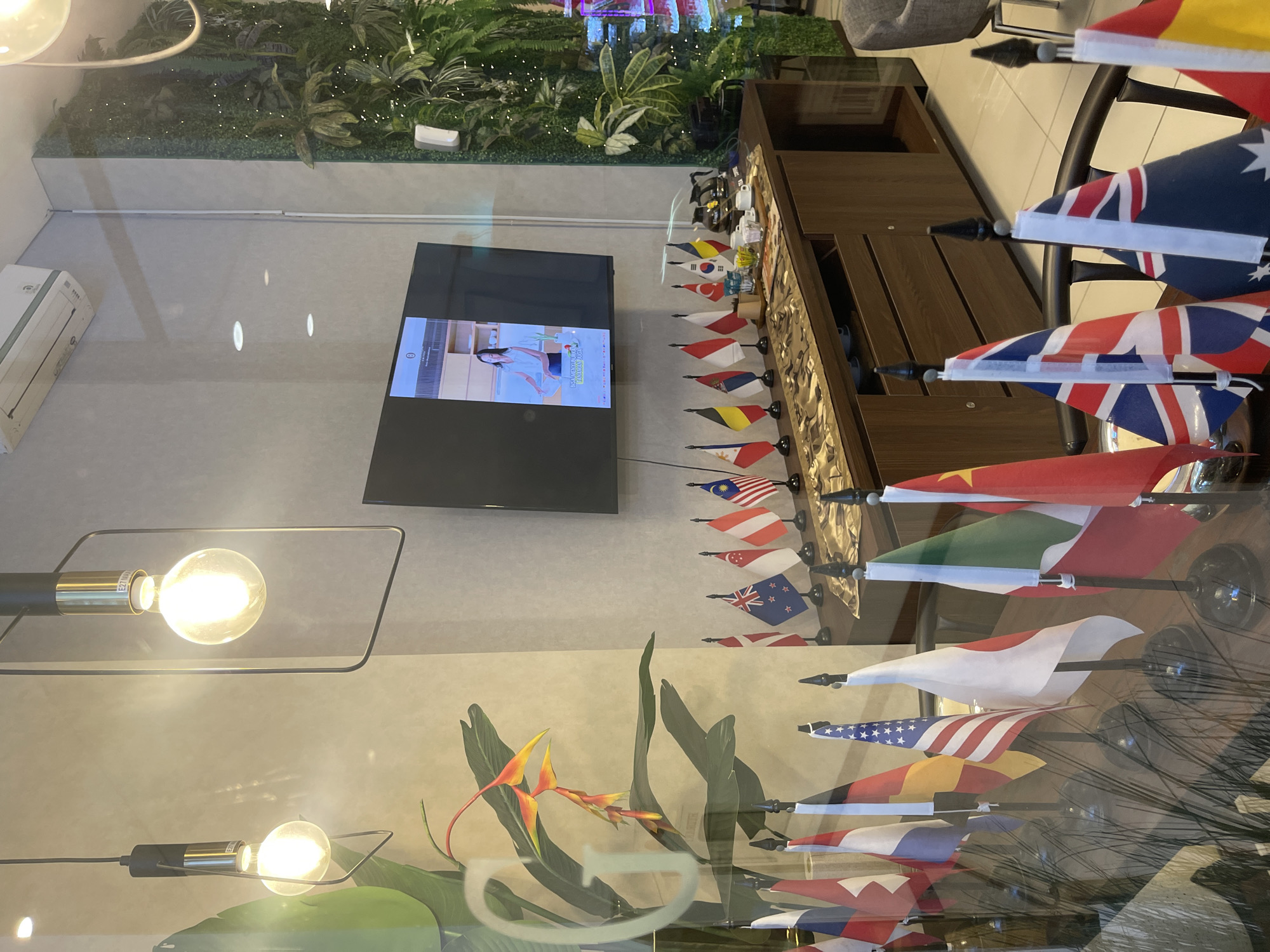 This media file captures an indoor scene, likely a modern office or commercial space, with a strong emphasis on international representation through a collection of small flags. The overall setting appears to be well-lit and contemporary, with reflections from glass surfaces suggesting it might be a storefront or an area with large windows.
The primary subjects are numerous small national flags displayed on stands, arranged in rows along a wooden counter or table. The flags represent a diverse range of countries, including Indonesia, the United States, Australia, the United Kingdom, China, and others that are partially visible. These flags are prominently featured, suggesting a context related to international business, diplomacy, or a globalized environment.
In the background, a large flat-screen television displays an image of a woman, possibly from a broadcast or a digital advertisement. To the left of the television, a white air conditioning unit is mounted on a light-colored wall. Above the counter and flags, a lush indoor garden or vertical planting installation is visible, adding a touch of nature to the otherwise modern interior. Two illuminated light bulbs, suspended from thin metal frames, are also noticeable, casting a warm glow.
The scene appears to be taking place indoors during the daytime, as indicated by the lighting and the reflections. There are no people actively engaged in a visible activity. The presence of multiple international flags suggests a connection to global affairs or a diverse clientele. The location context of South Jakarta, Indonesia, aligns with the Indonesian flag being among the displayed national symbols. No discernible text is clearly legible from the flags themselves, although the television screen might contain some text that is too small or blurry to read.