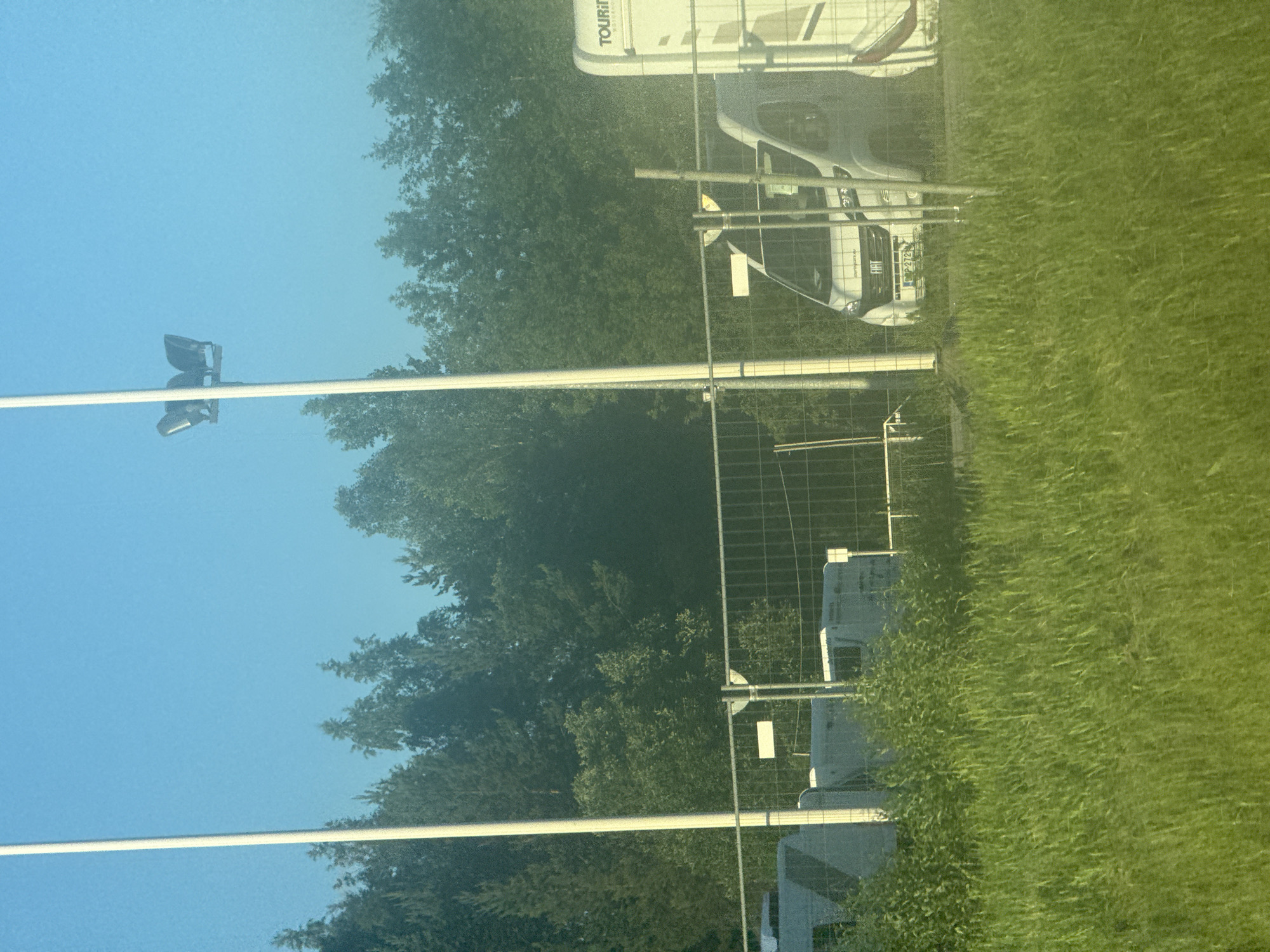 The image depicts a sunny outdoor scene with a clear blue sky. In the foreground, lush green grass is visible. A wire mesh fence runs horizontally across the middle of the frame. Behind the fence, several white camper vans and motorhomes are parked. One motorhome on the right has "TOURIN" visible on its side. A tall, thin white pole is prominent on the left side of the image, extending from bottom to top. In the upper portion of the frame, silhouetted against the sky, are three large stadium lights mounted on a pole. The background consists of a dense line of green trees. The vehicles appear to be stationary.