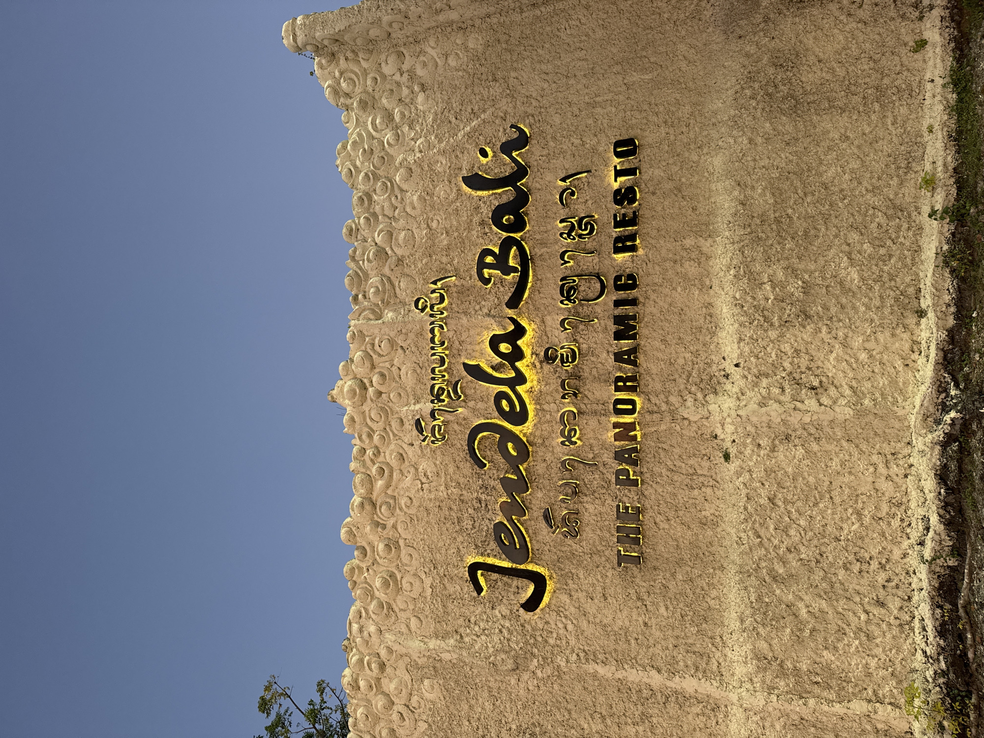 The image shows a large, textured wall with signage in Kuta Selatan, Indonesia. The wall is a light tan color and has a rough, somewhat bubbly texture across its surface. At the top of the wall, there is a decorative border with a repeating circular pattern. Above the wall, a clear blue sky dominates the upper portion of the image.
Prominently displayed on the wall are three lines of text. The top line is in a script that appears to be Balinese. The middle line, in a stylized, glowing yellow font, reads "Jendel Bali". Below this, a second line of Balinese script is visible. The bottom line, in a blocky, glowing yellow font, reads "THE PANORAMIC RESTO".
To the left of the wall, the branches and leaves of a tree are visible, extending into the upper left corner of the frame. The bottom of the image shows the ground, with some dry, white material along the base of the wall and green vegetation growing out from it. The overall impression is that of an entrance or facade for a restaurant.