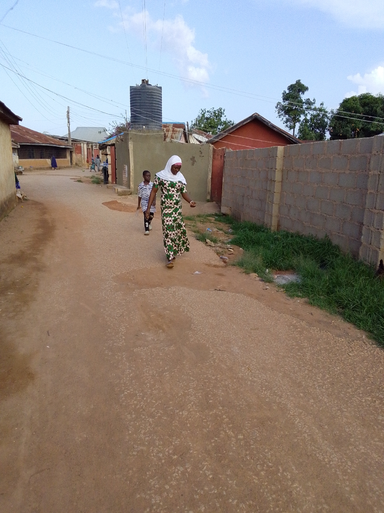 An eye-level, full shot captures a street scene in Jos, Nigeria, under a bright, partly cloudy sky. The setting appears to be a residential area with unpaved roads and a mix of traditional and modern housing.
In the foreground, a woman and a young boy walk along the light brown, sandy-gravel road, moving from the left side of the frame towards the right. The woman, positioned slightly ahead, wears a long, flowing green and white patterned dress (possibly ankara fabric) and a white hijab covering her head. She is looking forward and appears to be wearing sandals. Behind her and to her left, the boy wears a striped t-shirt (white and dark blue/black) and dark shorts, with the number "3" visible on his left leg. He is also looking forward and appears to be holding a small bottle or container in his left hand.
The road surface shows wear and tear, with patches of lighter gravel and dust. To the right of the road, a low, grey cinder block wall stretches into the distance, with patches of green grass and weeds growing at its base. Behind this wall, a house with a red roof is visible, surrounded by lush green trees.
On the left side of the street, various single-story houses with corrugated metal roofs line the road. Some appear to be older, possibly mud-plastered structures. Further down the road, additional figures, including a person in a blue garment, are visible near the houses. A large, black plastic water storage tank on a stand stands prominently in the mid-ground, surrounded by more houses and structures. Overhead, numerous utility lines crisscross the sky, connecting to poles that are partially visible. The overall impression is a bustling yet everyday scene in a local community.