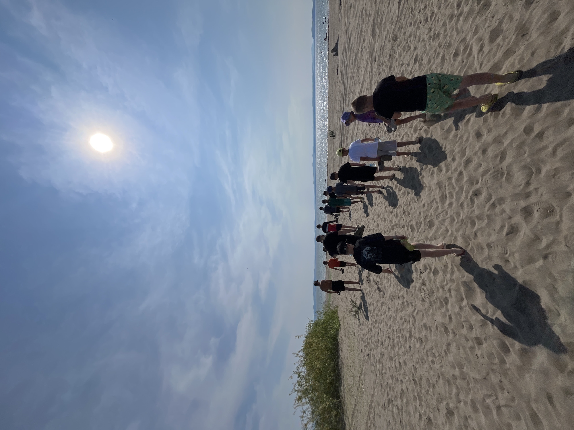A group of people are walking along a sandy beach. The sun is shining brightly in a clear blue sky with some wispy clouds. The ocean is visible in the distance, with mountains on the horizon. The scene appears to be set in the afternoon, given the length of the shadows cast by the people. The overall atmosphere seems relaxed and pleasant. The people are dressed in casual summer clothing, suggesting a warm day. There are no visible objects or text.