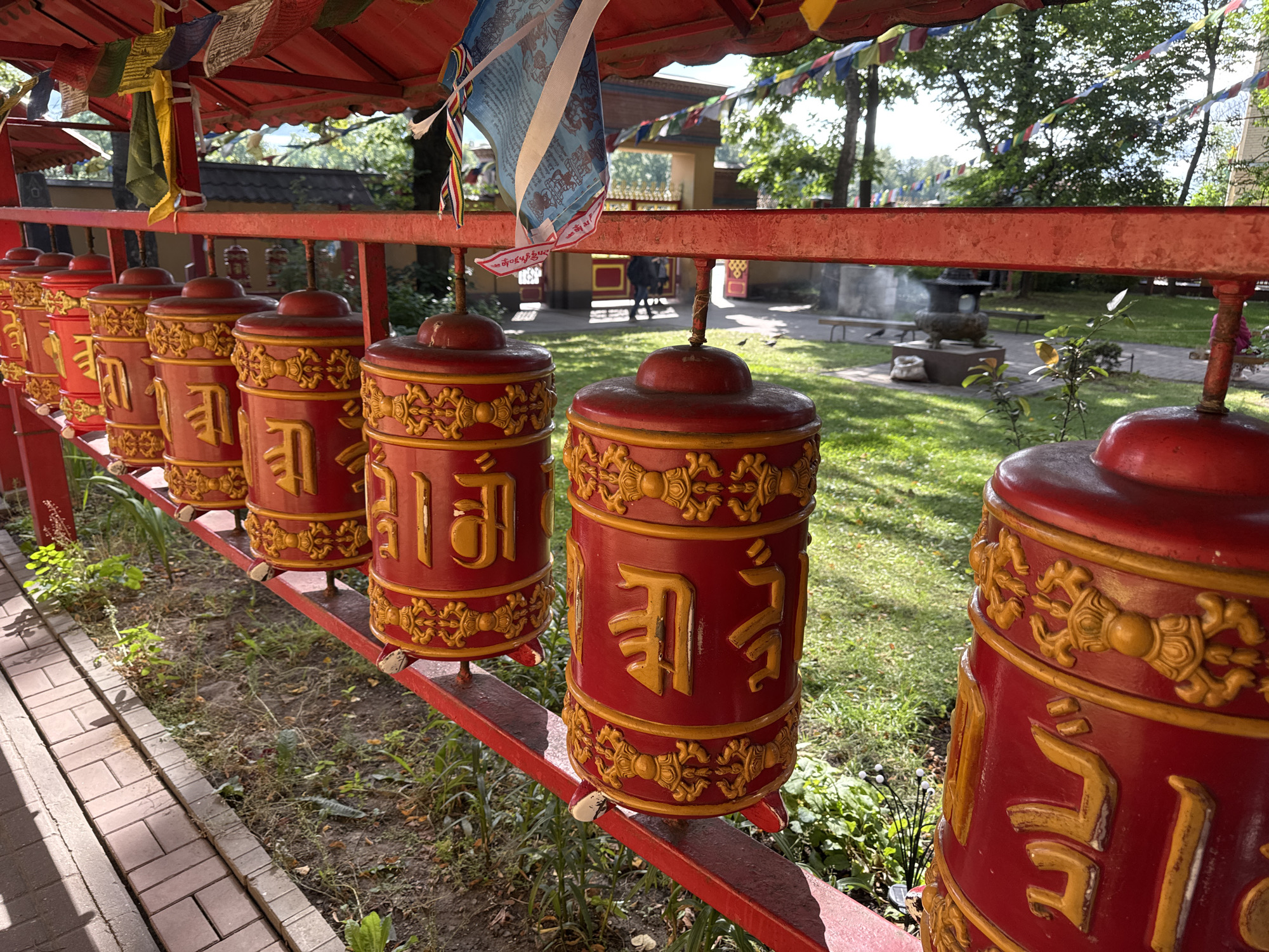 The image shows a row of red prayer wheels with golden Tibetan inscriptions, set on a red metal rack. They are located outdoors, in what appears to be a Buddhist temple complex. Prayer flags are strung above the prayer wheels, and greenery, including trees and grass, is visible in the background. A paved pathway is to the left of the prayer wheels. The scene is well-lit by sunlight. This appears to be the Buddhist Temple Datsan Gunzechoinei in Saint Petersburg, Russia, known for its distinctive architecture and religious artifacts.