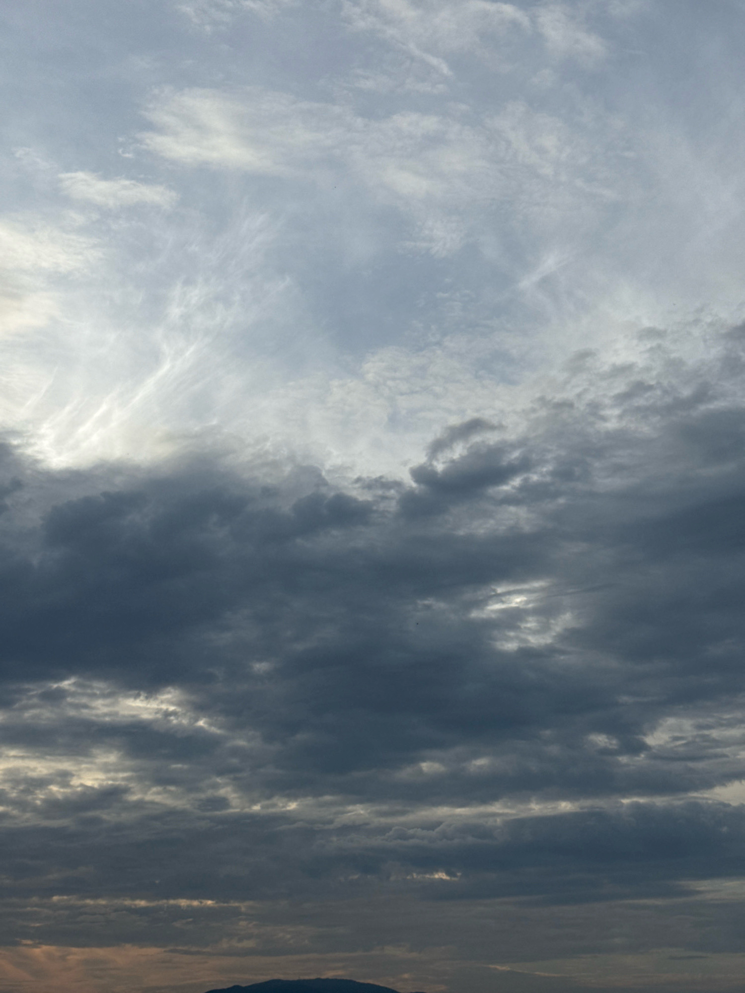 The image shows a cloudy sky over a distant dark silhouette of hills. The sky is filled with various cloud formations, ranging from wispy, thin clouds at the top to thick, dark, and heavy clouds lower down. The overall impression is of an overcast or pre-storm condition. The bottom of the image reveals a faint, dark, hilly horizon line, suggesting the photograph was taken from a vantage point with a view of distant terrain. There is no discernible indication of the city of Nha Trang, Vietnam, within the frame, as the focus is entirely on the sky and the distant horizon. The scene is devoid of people, specific objects, or any actions or interactions.