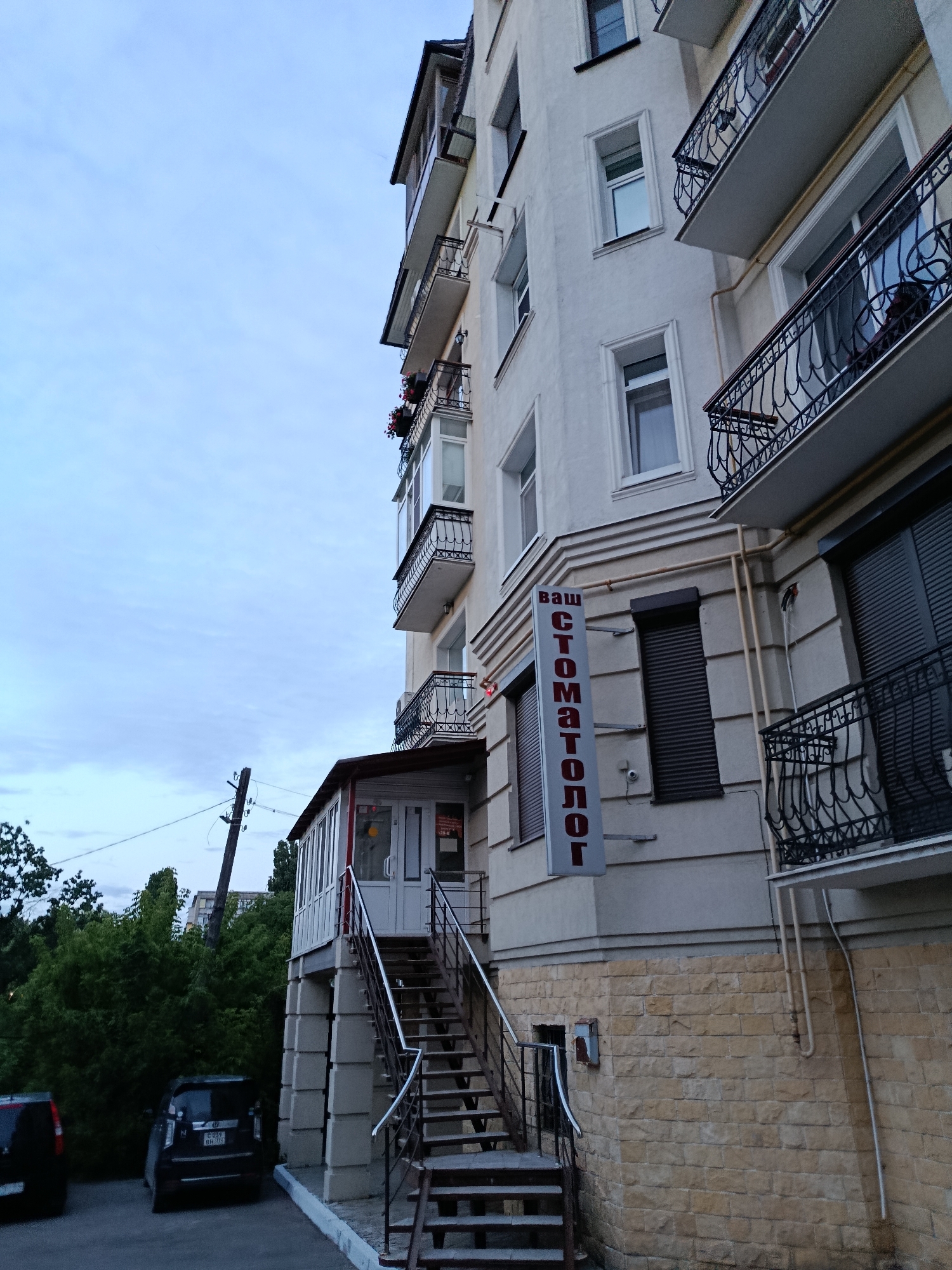 The image shows a view of a multi-story building from a low angle, looking upwards. The building is light-colored, with several balconies that have decorative metal railings. There is a sign on the side of the building with vertical red text that reads "ВАШ СТОМАТОЛОГ" (Your Dentist). Below the sign, there is a dark-colored roller shutter. A glass-enclosed entryway with a staircase leading up to it is visible on the lower level of the building. The entryway is adorned with red trim. To the left of the entryway, there is a black car parked, and behind it, a dark blue car is also parked. Behind the cars and the building's lower section, lush green trees are visible. A utility pole with wires extends upwards into the pale blue sky, which has a few wispy clouds. The overall scene depicts the exterior of a building in an urban environment, likely a residential or mixed-use area. The specific location is identified as Saratov, Russia.