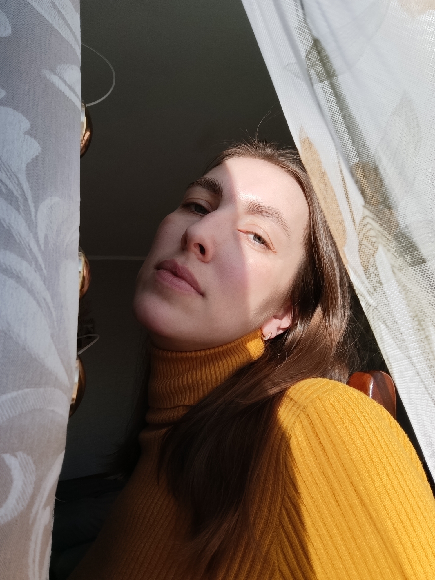 A young woman with light skin and brown hair, wearing a yellow turtleneck sweater, is positioned in front of a window. Part of her face is illuminated by sunlight. She is looking slightly upwards and towards the camera. The window is covered by a white patterned curtain. A dark interior is visible to the left of the curtain, with a gold-colored object partially visible. A part of a wooden chair is also noticeable. The location cannot be determined as being in Фоки, Russia from the image alone.