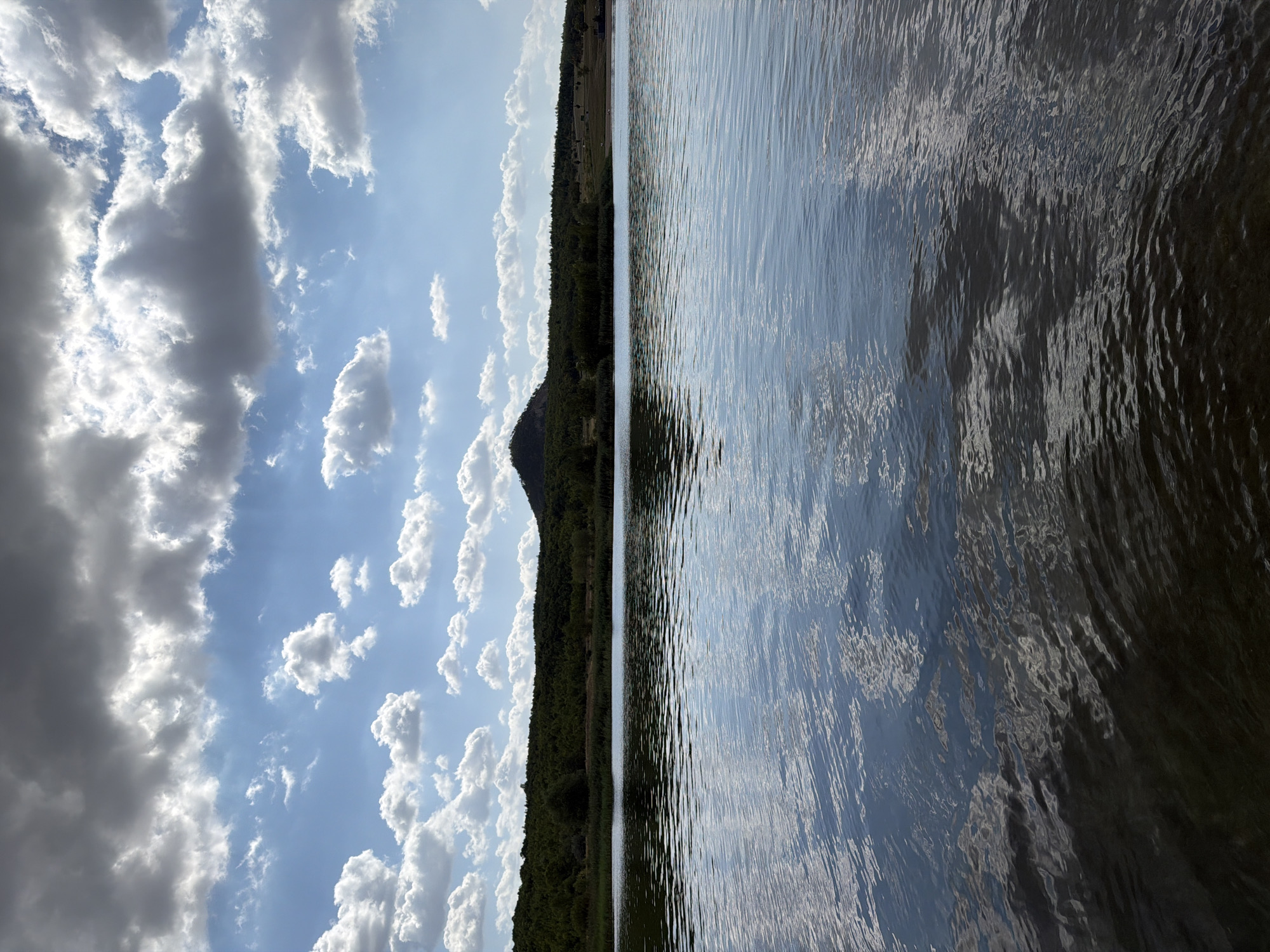 A serene lake reflects a dramatic sky filled with dark and light clouds. A tree-lined hill with a small peak dominates the horizon, suggesting a natural landscape. The water is calm, with gentle ripples disturbing the surface, creating abstract patterns of light and shadow. The scene is devoid of any human presence or man-made structures, evoking a sense of untouched nature. It is highly probable that this image depicts the water body known as the water reservoir Bedřichov, located near the city of Most in the Czech Republic.