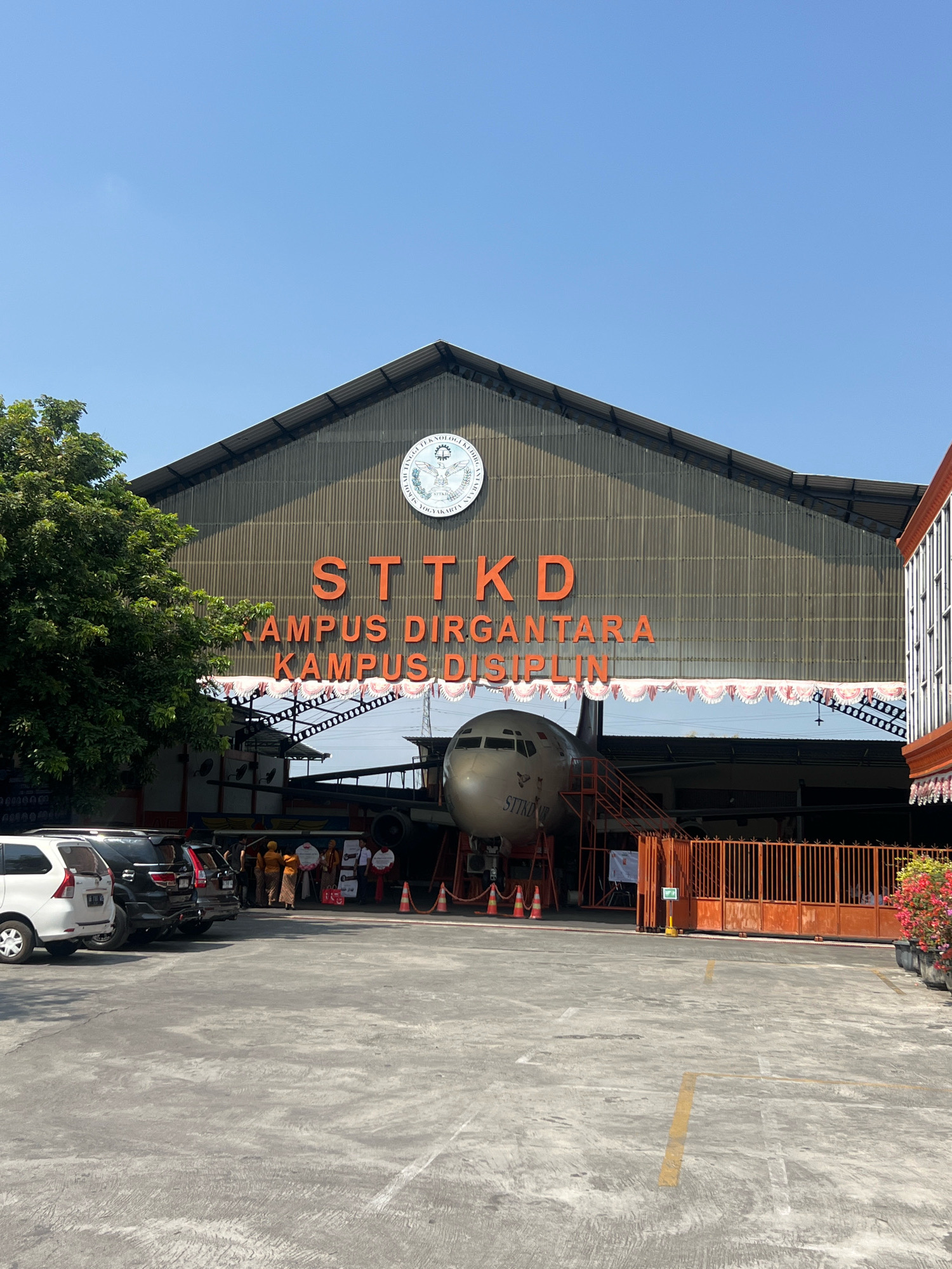 The image captures an outdoor scene in front of a large hangar-like building, identified as the STTKD (Sekolah Tinggi Teknologi Kridha Budi Bhakti) campus in Sewon, Indonesia. The building's facade features the prominent orange text "STTKD KAMPUS DIRGANTARA KAMPUS DISIPLIN". Centered within the open hangar doors is the nose and front section of a silver airplane, its registration number "STTKD" visible on the fuselage. Red stairs lead up to the airplane's door.
In the foreground, a paved parking lot is visible with several cars parked on the left. Several people are gathered in the middle of the scene, some wearing traditional Indonesian attire. Orange traffic cones are placed on the asphalt. To the right, an orange metal fence encloses an area, and a lush bush with red flowers is in the far right corner. The sky is clear and blue. The overall atmosphere suggests an institutional setting, possibly for an event or a demonstration related to aviation technology.