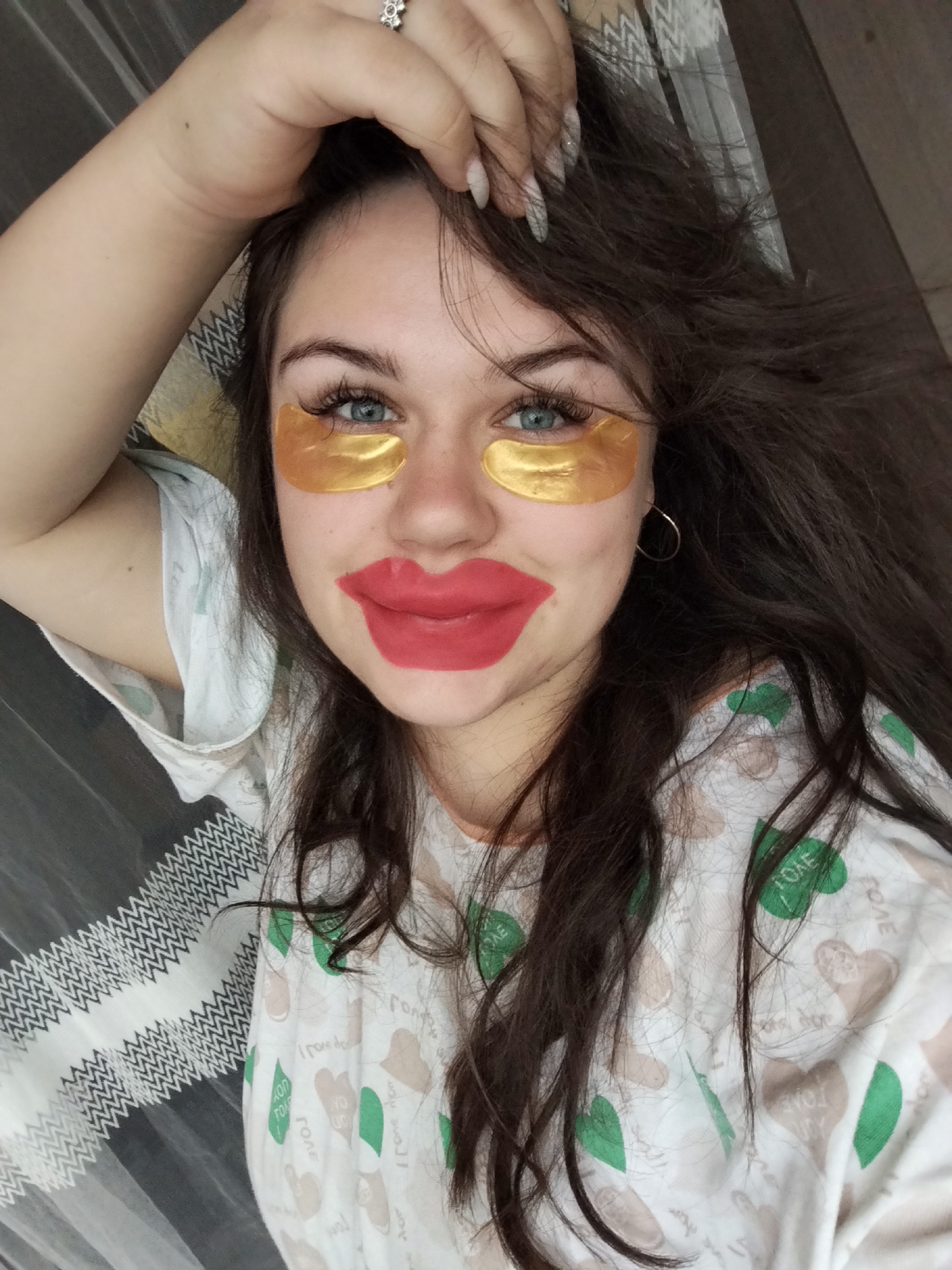 A young woman is depicted indoors in what appears to be a private residence in the city of Chaikovsky, Russia. She is lying down with her head resting on a bed or a similar surface covered with a patterned sheet. She is wearing a white shirt with a pattern of green hearts and beige heart shapes with the word "LOVE" on them. She has brown hair and blue eyes. She is wearing gold eye patches under her eyes. A red, oversized lip mask covers her lips and the skin immediately around them. Her left hand is raised and positioned near her forehead, and she is wearing a silver ring on her left ring finger. The overall lighting in the room is soft and diffused.