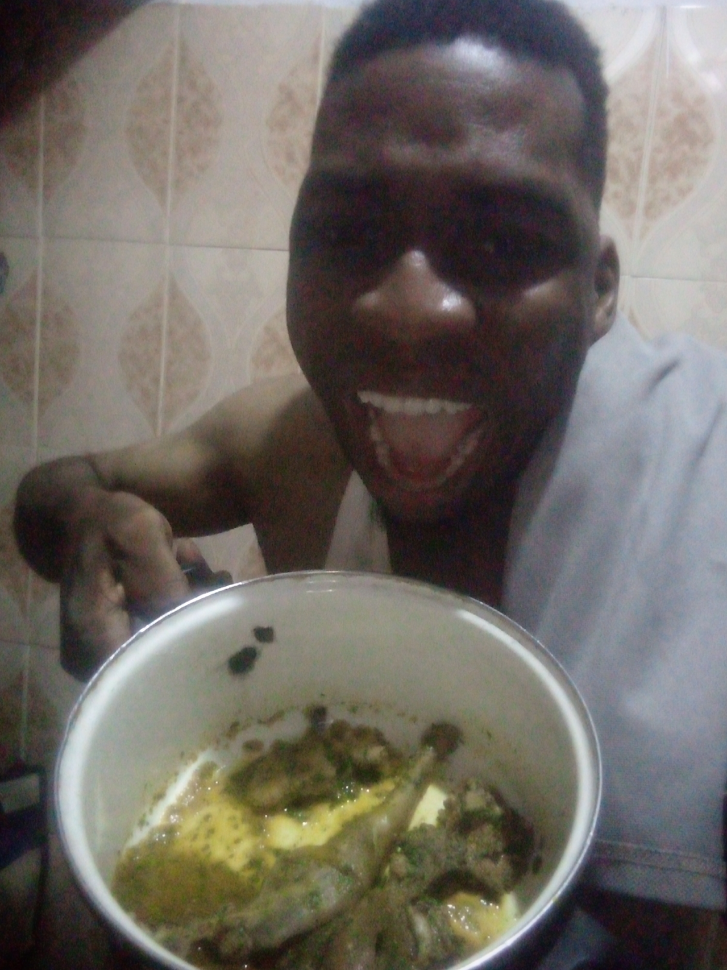 A close-up, waist-up photograph depicts a smiling, dark-skinned person holding a pot of food. The person has short, dark hair, visible teeth, and is wearing a light-colored garment. The pot, white with a silver rim, contains a stew-like dish with pieces of meat and a greenish-yellow sauce. The background features a wall with patterned beige tiles. The location appears to be a bathroom, but the city and region are unknown.