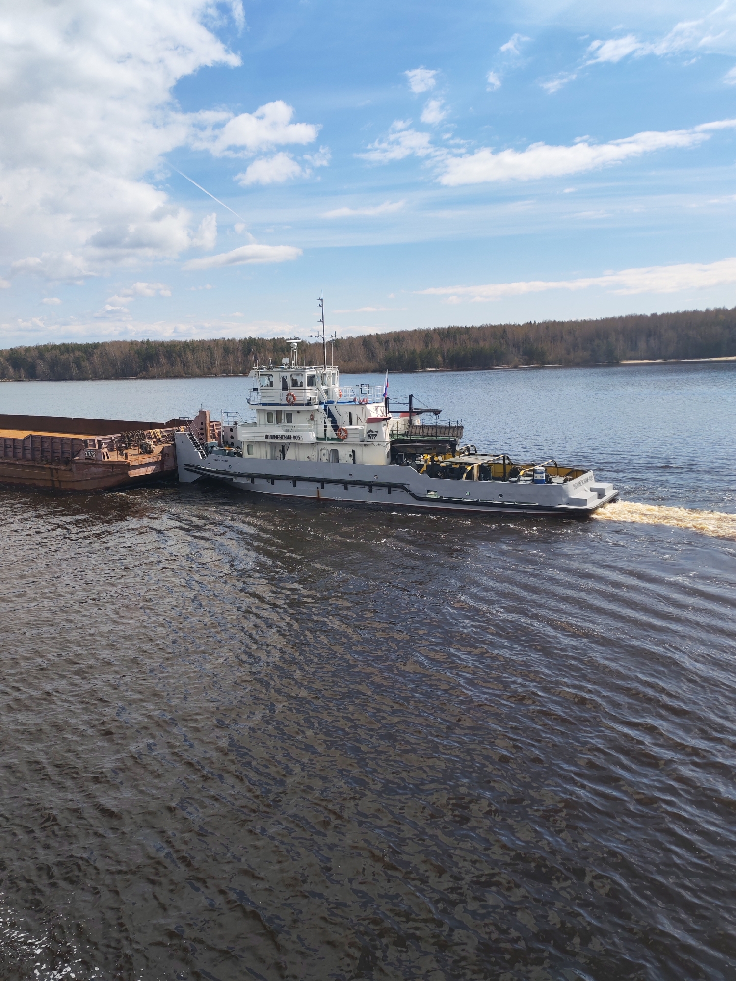 A tugboat, labeled "КОЛОМЕНСКИИ-615", is moving on a body of water, pulling a barge to its left. The water is dark and rippled, with a visible wake behind the tugboat. The shoreline is visible in the background with trees. The sky is blue with scattered clouds and a contrail. The location and specific city are unknown.