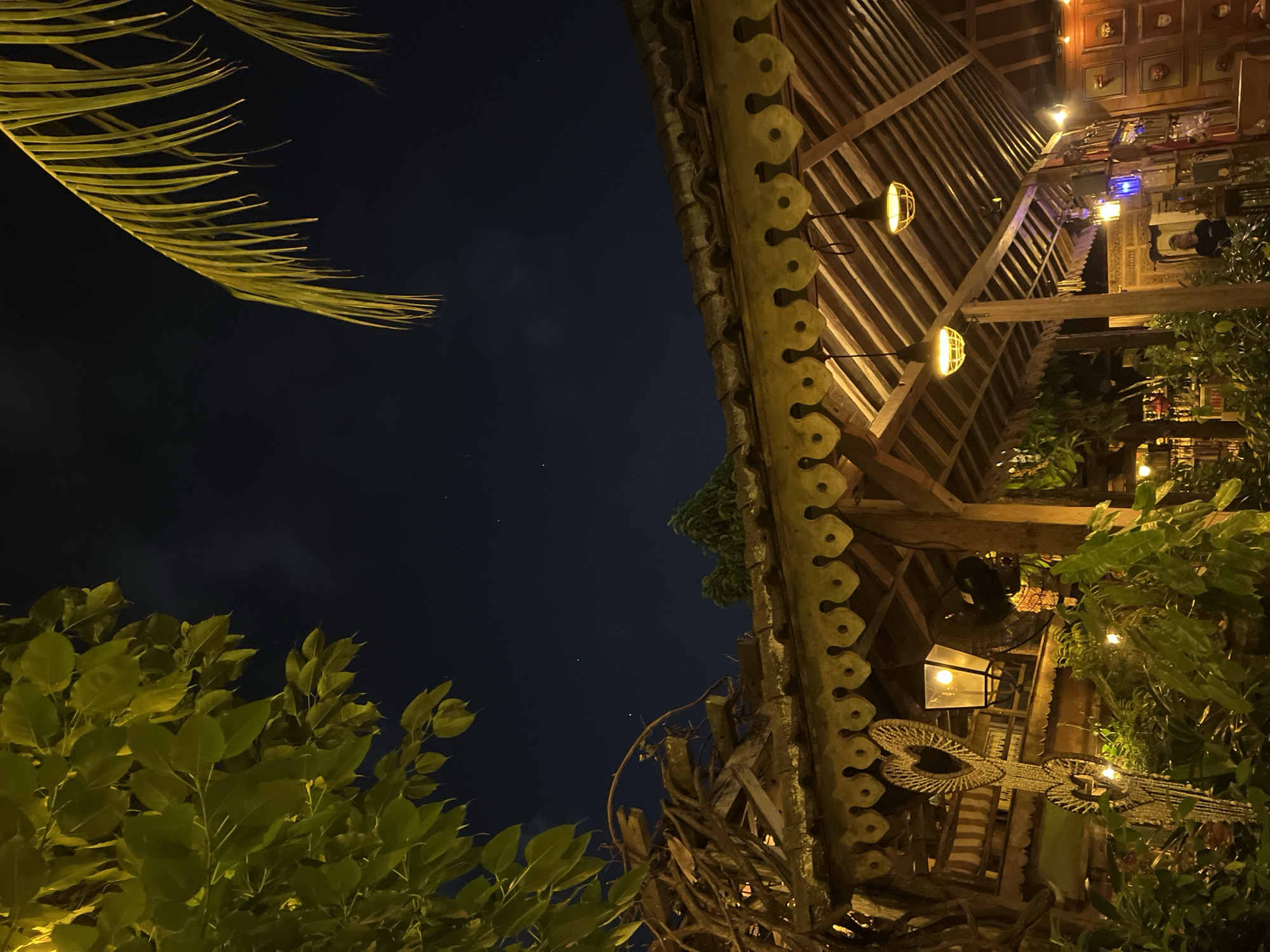 Captured at night in North Kuta, Indonesia, the image presents a low-angle shot toward a structure, possibly a building or gazebo. A dark night sky occupies the upper left corner, showing a few stars and some cloud cover. The structure is predominantly constructed of wood, featuring a distinctive roofline with decorative trim, round pendant lighting, and other ornate details. Abundant foliage frames the composition, with green leaves and palm fronds in the foreground, enhancing the tropical atmosphere. A light fixture with a lamp shines dimly. Overall, the scene conveys an ambiance of a cozy, illuminated space set amidst a verdant tropical environment at night.