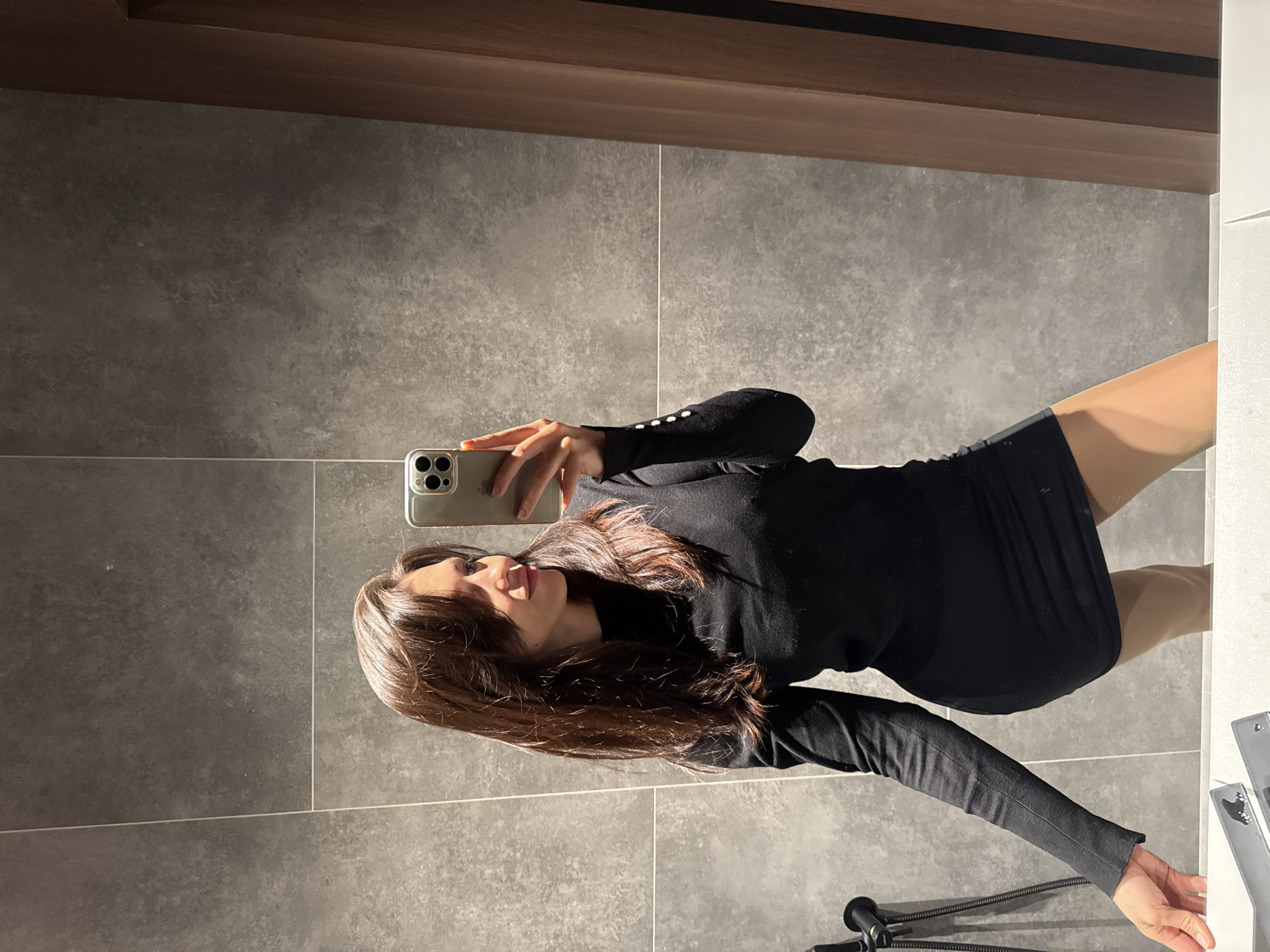 A woman in a black turtleneck sweater and skirt takes a selfie in a bathroom with grey tile walls and a brown cabinet. The location is unknown.