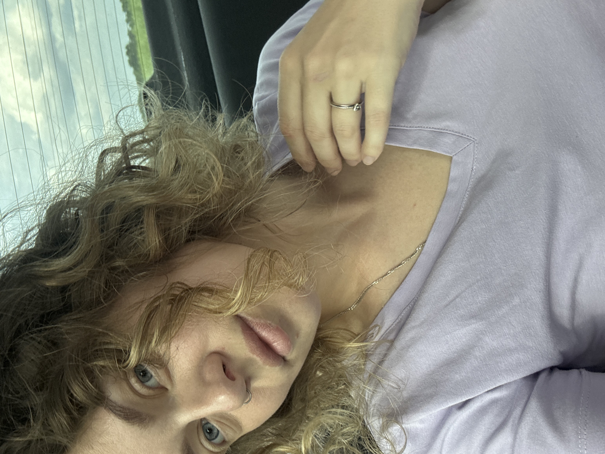 This is a close-up, selfie-style portrait of a young woman with blonde, curly hair, blue eyes, and a nose ring. She is wearing a light purple V-neck t-shirt and a silver necklace. A silver ring is visible on her right hand, which is resting on her neck. The background appears to be the interior of a vehicle, with a car window showing a cloudy sky and a green, distant landscape. The setting does not provide any specific information about the city of Nadezhda, Russia.