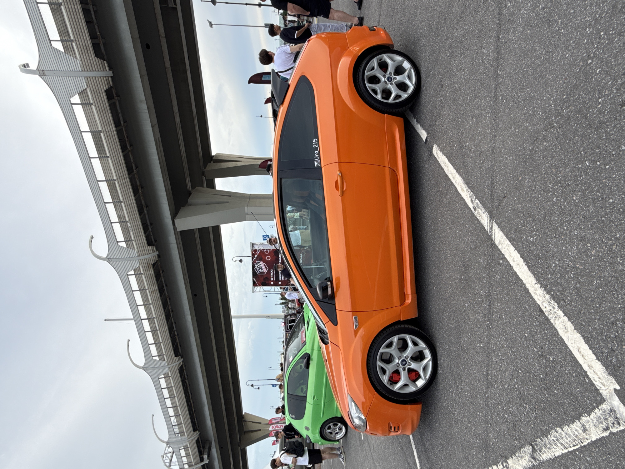 An outdoor event is taking place under a large, modern overpass in Saint Petersburg, Russia. The focus is on a vibrant orange Ford Focus ST parked on an asphalt lot marked with white lines. The car features distinctive silver alloy wheels with red brake calipers. In the background, a bright green Peugeot 207 is also visible, along with several people standing around the vehicles. A large advertising banner, possibly for a car-related event, is mounted on a scaffold structure behind the cars. The sky is overcast, suggesting a cloudy day. The overpass, a prominent architectural feature, appears to be part of a major highway or urban expressway system.