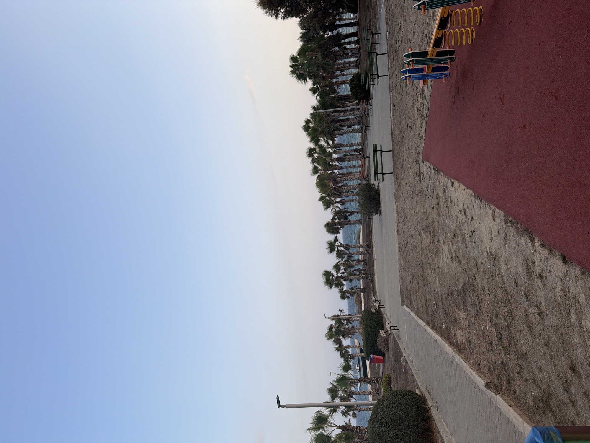 The image shows a seaside park in Pervolia, Cyprus, at what appears to be dusk or early morning. The sky is a clear, pale blue, with a faint line of clouds near the horizon. A row of palm trees lines the edge of a body of water, likely the Mediterranean Sea, which is calm and exhibits a deep blue color. A light-colored boat is visible on the water in the distance.
In the foreground, a paved walkway runs diagonally from the lower left to the mid-right of the image. To the left of the walkway, there are manicured bushes and a lamppost. Further along the walkway, several empty park benches are situated, facing the sea and the palm trees. A red trash can is also visible near the walkway.
The ground in the park is a mix of dry, sandy soil with sparse vegetation. In the lower right quadrant of the image, a portion of a red playground surface is visible, with a spring rider playground toy featuring yellow springs and blue and green seats. The scene is peaceful and empty, suggesting a quiet time in the park.