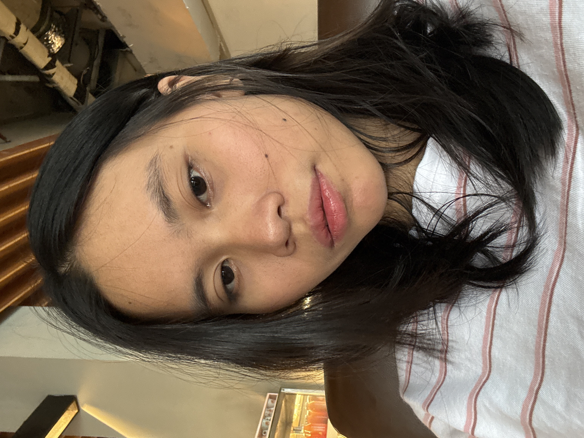 A close-up, overhead, selfie-style shot shows a young East Asian woman with long, dark hair, wearing a white t-shirt with thin pink and brown stripes. Her face is turned slightly to her left, and she is looking directly at the camera with a neutral expression. Her skin is clear with a few small blemishes. A mole is visible on her left cheek. Her lips are slightly parted and appear to have a light sheen of lip gloss. The background shows a dimly lit interior with what appears to be a display case with bottles of beverages on the left, and industrial piping and wooden beams on the ceiling in the upper right portion of the frame. The overall lighting is warm and somewhat diffused. The image is taken in East Jakarta, Indonesia.