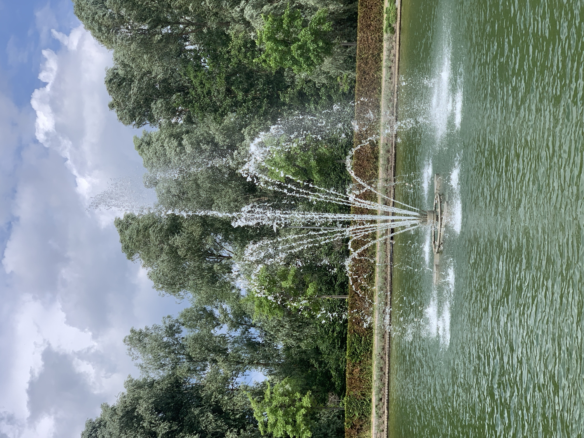 The image depicts a fountain in a pond, surrounded by lush greenery and trees. The pond water is a vibrant green, with ripples and splashes from the fountain. The fountain itself is a multi-tiered structure with water spraying upwards and outwards in a fan-like pattern. Behind the pond, a well-maintained hedge forms a boundary, followed by a dense line of trees with light green foliage. The sky above is partly cloudy, with a mix of white and grey clouds against a blue background. The scene appears to be a tranquil park or garden setting, likely in a temperate climate. The location is identified as being in the city of Bucha, Ukraine.