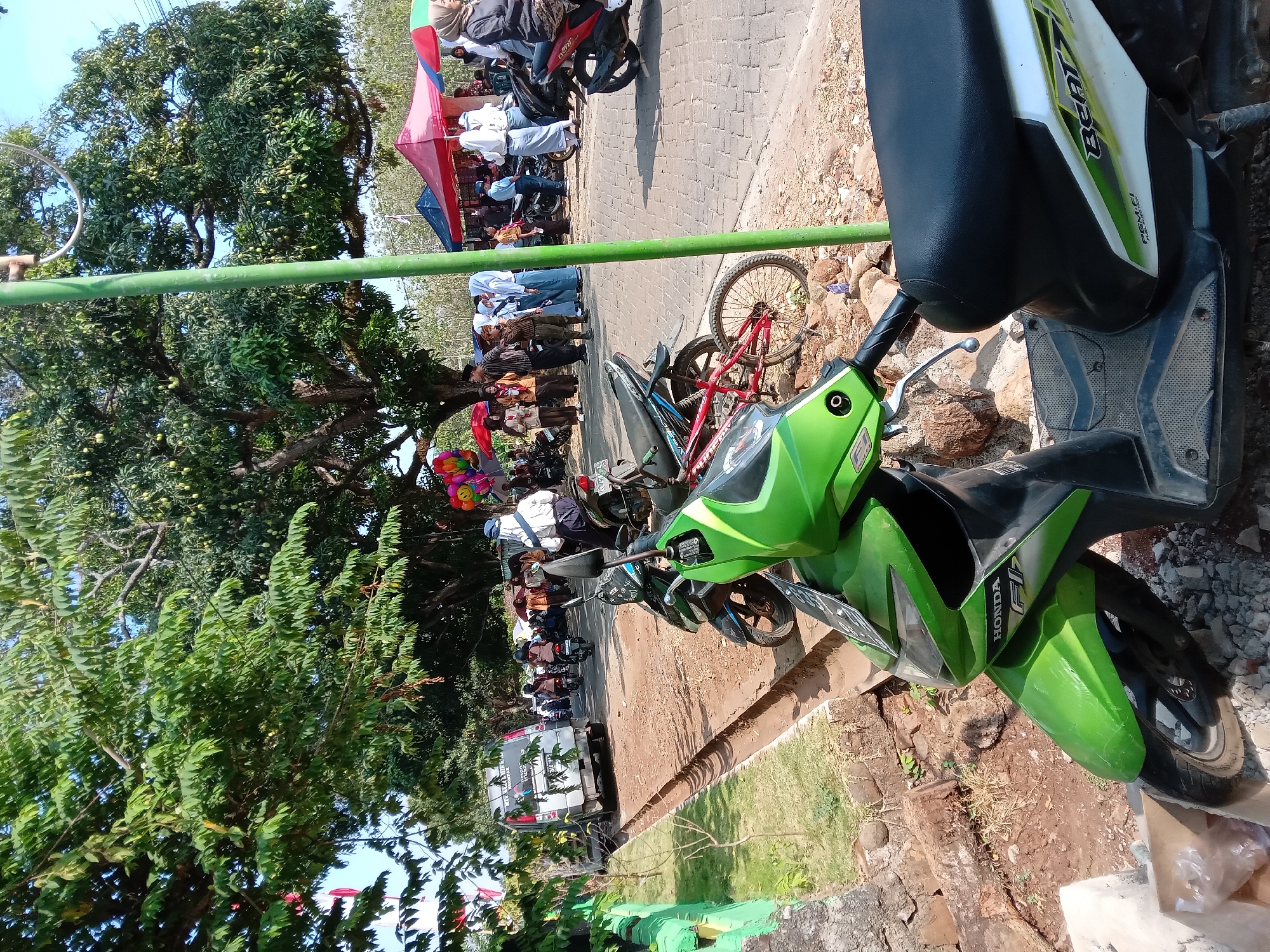 The image depicts an outdoor scene on a sunny day, likely in Ngrambe, Indonesia, given the context. In the foreground, a bright green and black scooter is parked on a dirt and gravel area next to a roadside trench. Behind the scooter, a red bicycle is also visible. Further into the scene, a paved road is lined with several motorcycles and a silver van. A group of people, some in uniforms (possibly students), are gathered near red and blue tents, suggesting a market or event. Tall trees with lush green foliage dominate the background, providing shade. A light pole stands prominently in the middle ground. The overall impression is of a bustling local gathering in a somewhat unpaved area adjacent to a road.